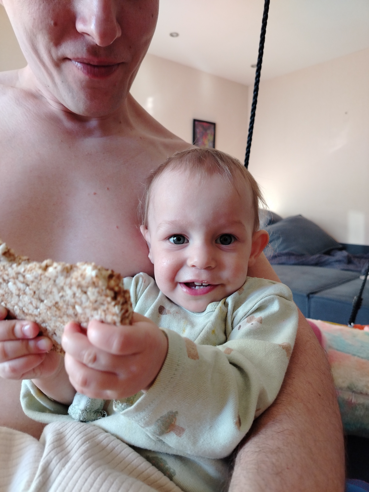 A man holds a baby in his arms. The baby is holding a piece of bread. The location is not specified in the image. The man is topless. The baby is wearing a long-sleeved shirt. There is a sofa in the background. A black cord is visible in the upper right corner. There's a picture frame hanging on the wall.