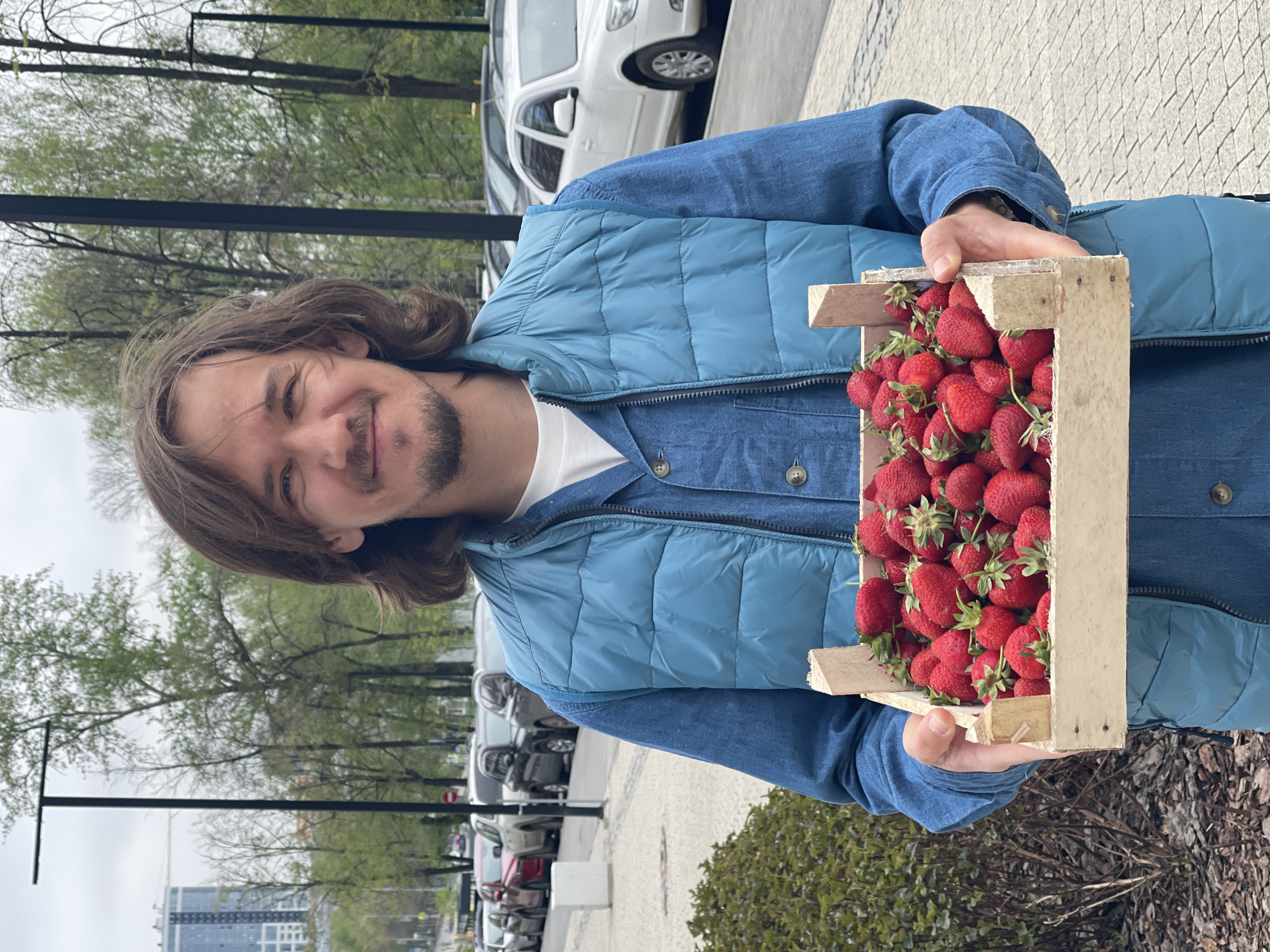 A man is holding a wooden crate filled with strawberries. He has long brown hair and a light beard and mustache. He is wearing a blue denim jacket and a light blue vest over a white t-shirt. The crate is full of bright red strawberries with green stems and leaves. The background shows a city street with trees, parked cars, and a building in the distance. The photo was taken in Izhevsk, Russia.