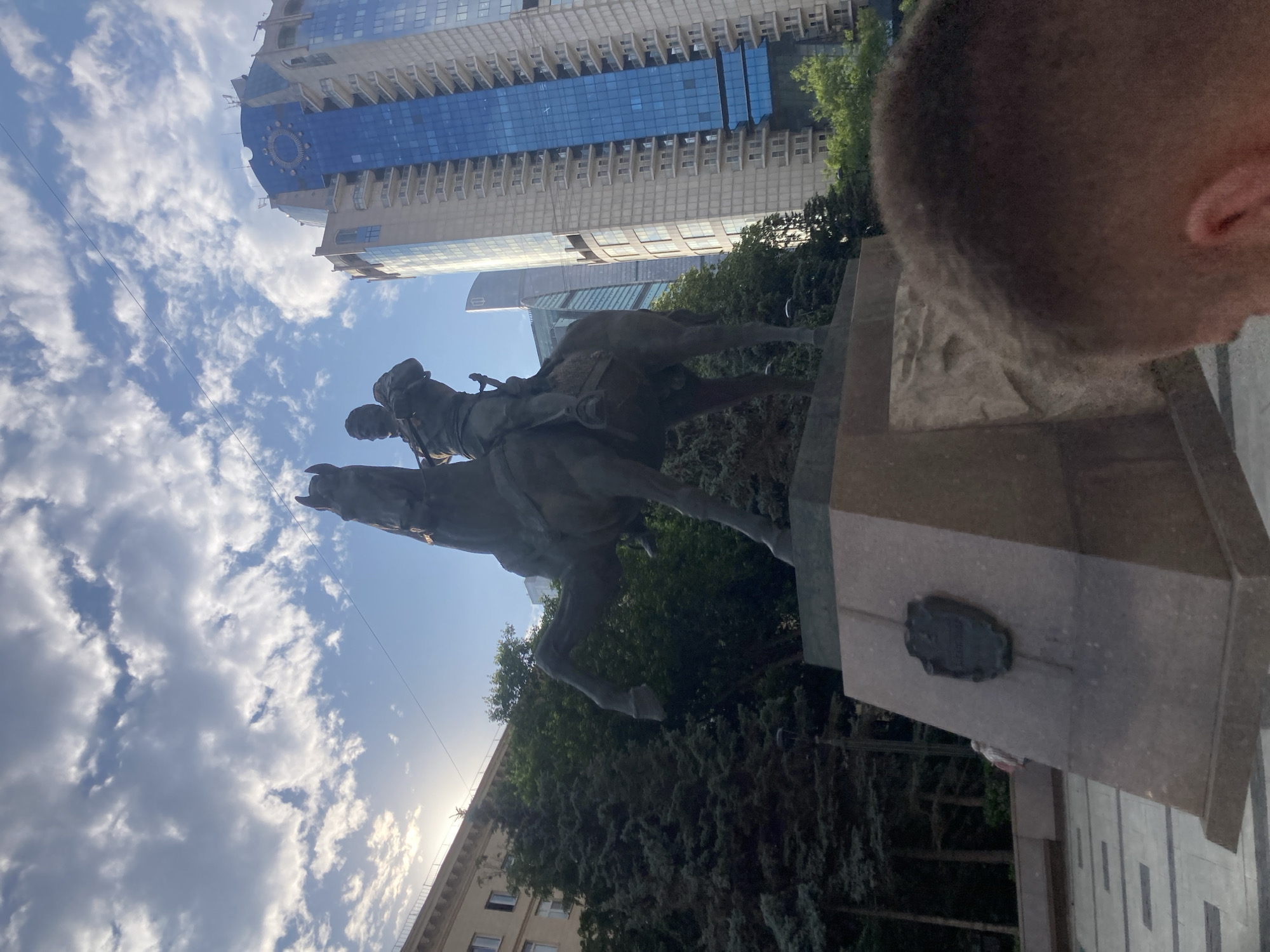 The image shows an equestrian statue in a city square, likely in Moscow, Russia, given the architecture of the surrounding buildings. The statue depicts a man in military attire on a rearing horse. The base of the statue has a plaque with an inscription, though it is unreadable due to blurriness and distance. In the background, a modern high-rise building with a distinctive blue facade and a circular clock-like emblem is visible. Trees and other buildings, including what appear to be skyscrapers, are also part of the urban landscape. The sky is partly cloudy with the sun peeking through. A person's head and shoulders are in the foreground, partially obscuring the bottom right of the statue. The ground in front of the statue is paved with stone tiles.