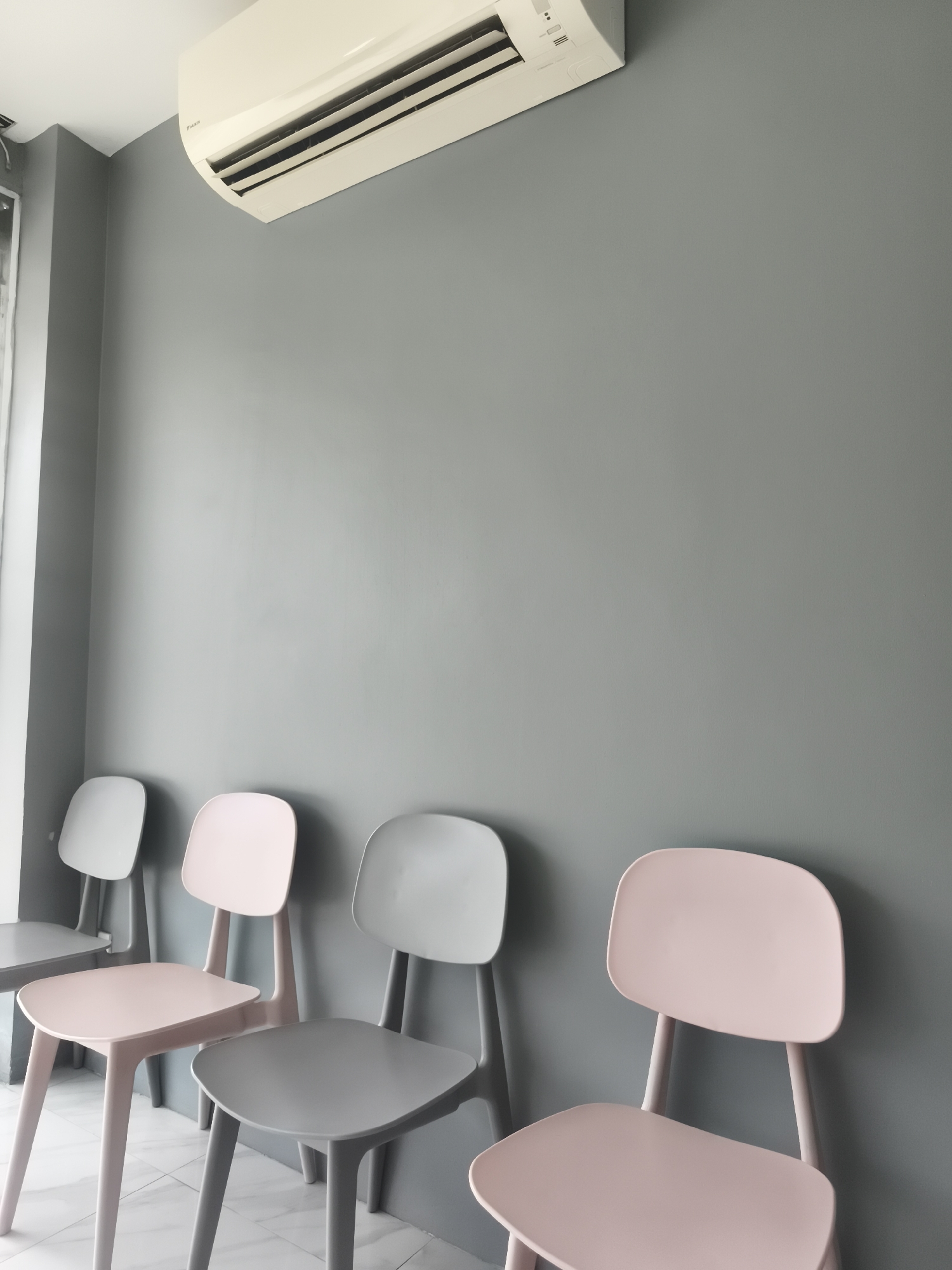 A row of chairs, some light pink and some grey, are lined up against a grey wall. An air conditioner unit is mounted on the wall above the chairs. The room appears to be a waiting area, possibly in a clinic or office. The floor is tiled with a light-colored marble pattern. There are no people visible in the image. The lighting is soft and even, suggesting an indoor setting with artificial light.