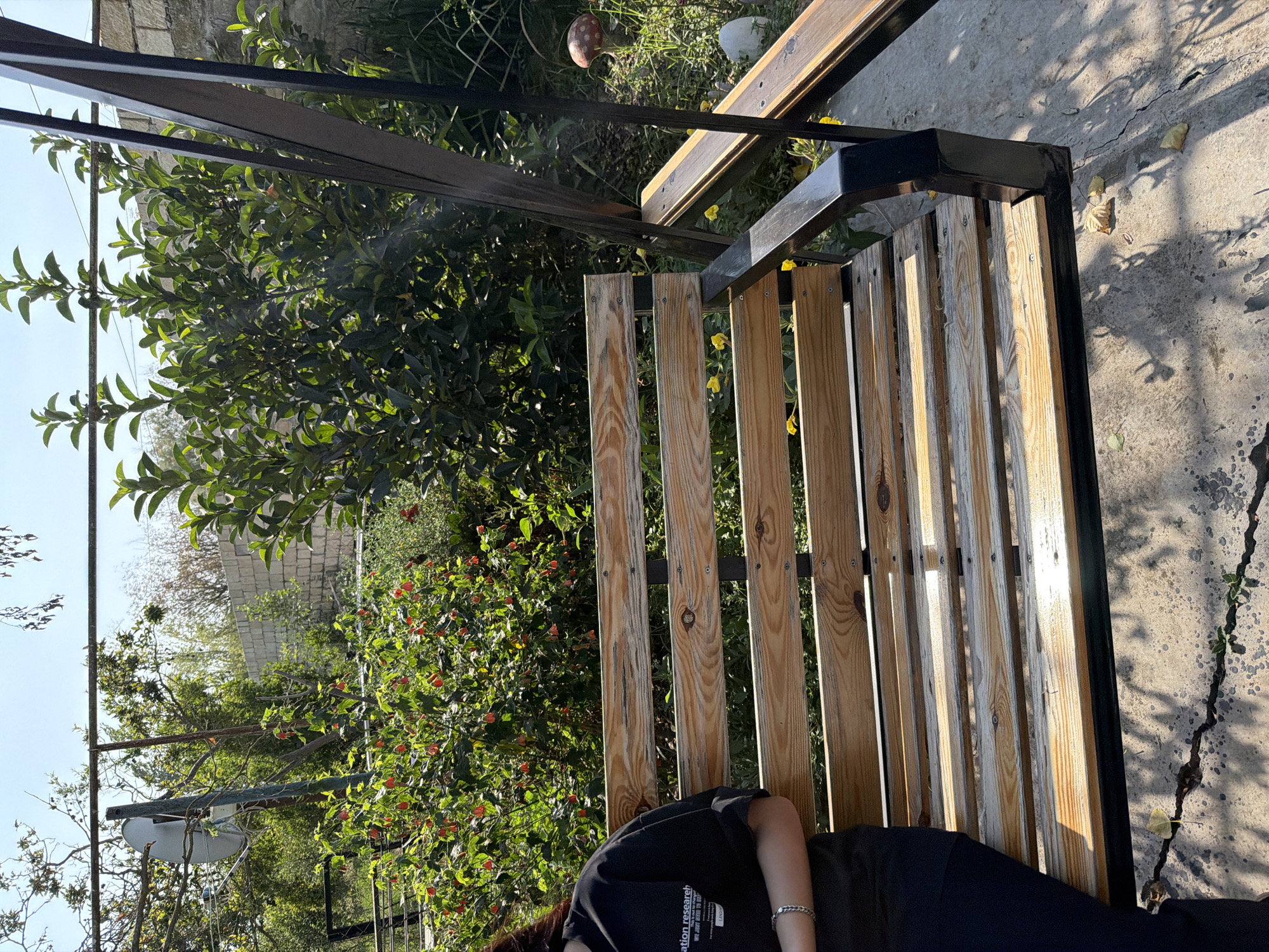 The image shows a wooden swing set with a person's torso and legs visible, sitting on the swing. The swing has a metal frame. The background consists of lush green trees and bushes, a brick wall, and a bright sky. There is a satellite dish mounted on a pole in the upper left portion of the image. A decorative mushroom figure is visible to the right of the swing. The ground beneath the swing appears to be concrete with some cracks. The overall setting suggests an outdoor garden or yard area. The location is specified as Balakənd, Azerbaijan.