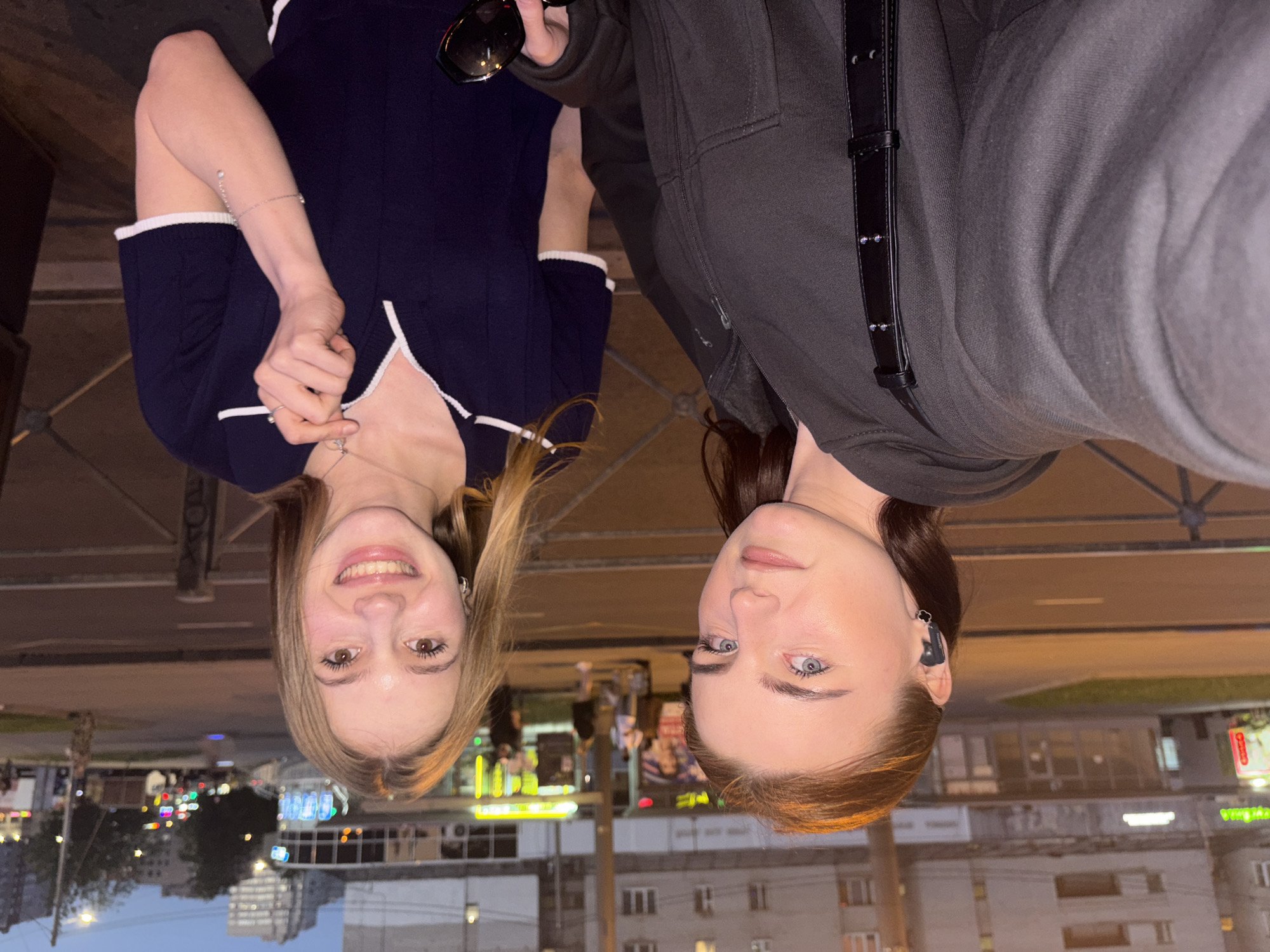 The image displays a selfie-style photograph taken at twilight or early evening on an urban street in Saint Petersburg, Russia. Two young women are positioned in the foreground, with one holding the camera.
The woman on the left has fair skin, dark brown hair, and blue eyes, maintaining a neutral expression while looking directly at the camera. She wears a dark grey hooded zipper jacket and a black strap from a bag, adorned with small silver studs, crosses her right shoulder. A dark earbud is visible in her left ear, along with a small black earring.
The woman on the right has fair skin, light brown hair, and brown eyes, smiling broadly at the camera. She wears a dark blue short-sleeved polo shirt with a white collar and white trim on the sleeves. A thin silver-colored chain necklace with a small pendant rests on her chest, and a delicate silver-colored bracelet with a single charm hangs from her right wrist. She holds dark sunglasses in her left hand, while her right hand is positioned near her necklace.
Behind the two individuals, a dark metal barrier or railing is visible. Beyond this, a paved street with white lane markings stretches into the mid-ground. Buildings line the street, characterized by multi-story residential or commercial structures. On the left, an older, grey building features visible windows. Towards the center and right, commercial storefronts are brightly lit, displaying various signs. One notable sign on the right side of the street features Cyrillic text, reading "Твоя" (Tvoya) in bright yellow letters. Other illuminated signs and streetlights are scattered throughout the background. The sky above is a deep blue, indicative of the evening hours, with distant building silhouettes. The overall scene captures an active urban environment.