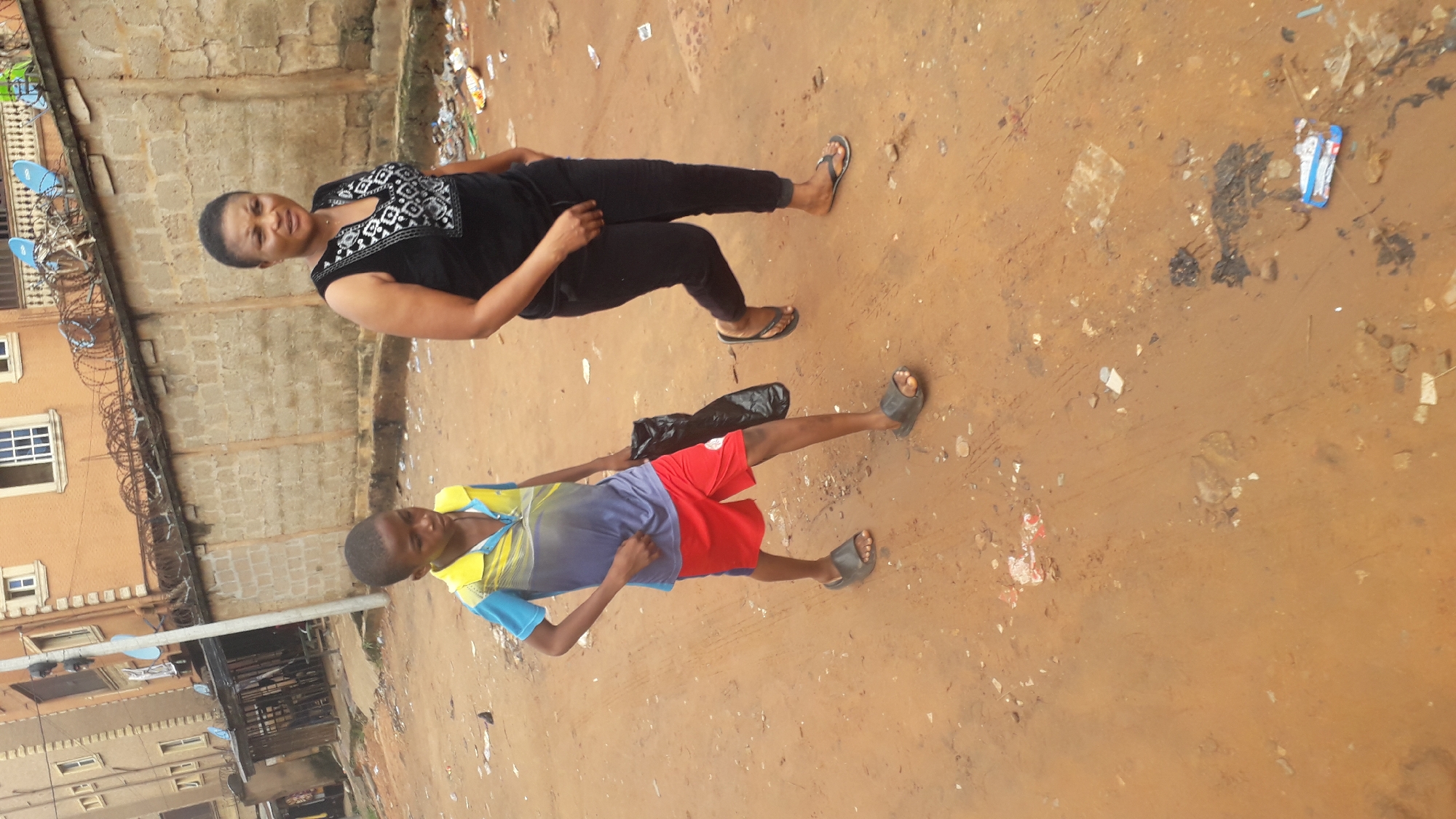 Two people are standing in an unpaved, debris-strewn area in Nkpor, Nigeria. An adult woman is wearing a black top with a white pattern, black pants, and sandals. A young boy is wearing a blue, yellow, and white shirt with red shorts and sandals, and is holding a black plastic bag. The background includes a building with barbed wire and light-colored brick and plaster. There is trash and debris scattered throughout the area.