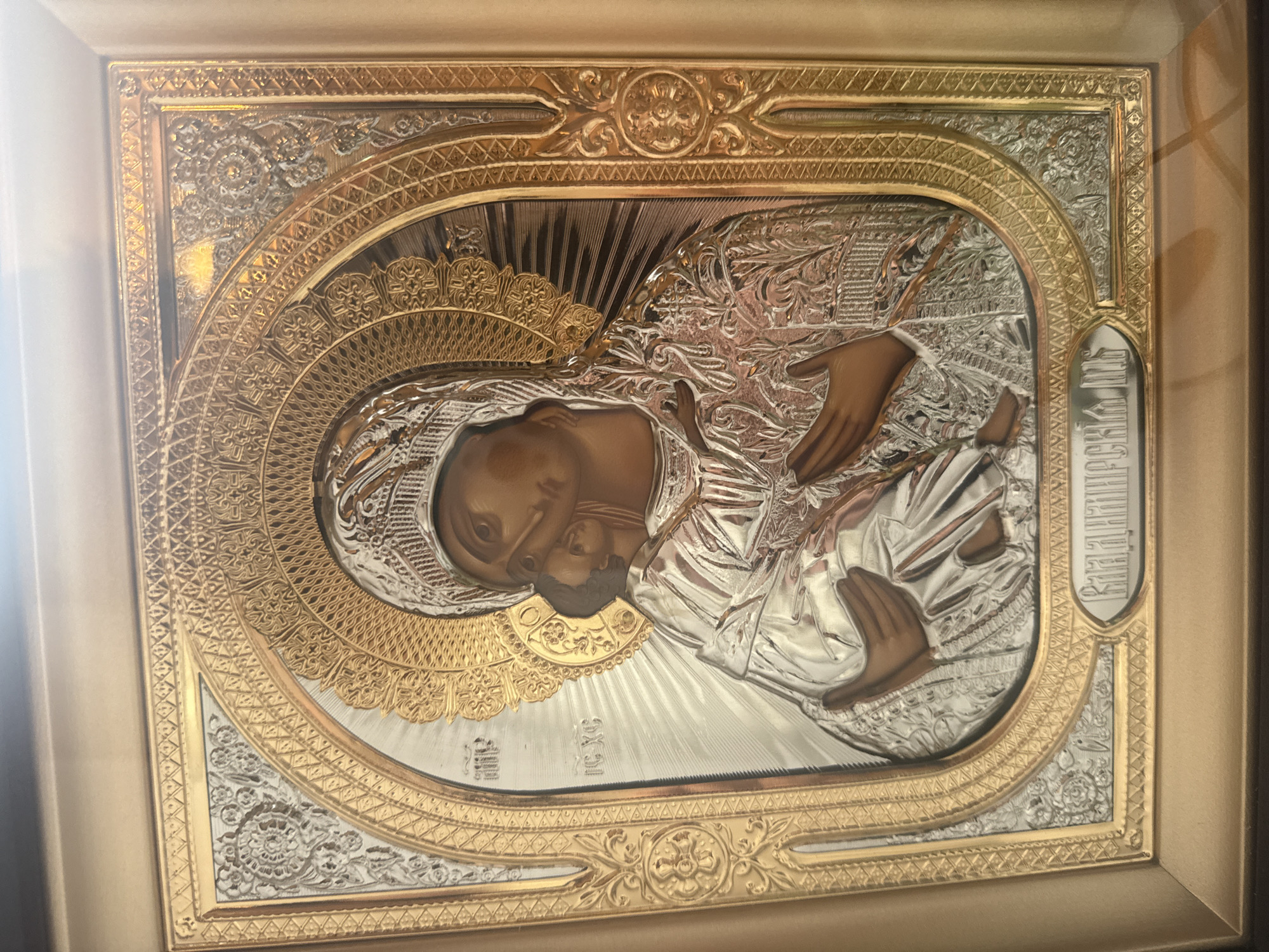 The media file displays a framed religious icon, likely of the Virgin Mary and Christ Child, rendered in embossed metal with gold and silver finishes. The central figures are depicted in profile, with the Virgin Mary embracing the child. Their faces and hands are rendered in a darker, flesh-toned metal, contrasting with the ornate, highly detailed silver and gold metallic robes and halos. The background behind the figures features radiating lines, suggesting a divine light. The icon is encased in a decorative frame, with intricate gold detailing on the inner border. The outer frame is a simpler, golden rectangular border. The overall aesthetic suggests a traditional Orthodox Christian icon, possibly a representation of the Hodegetria icon type. There is some Cyrillic text visible vertically on the right side of the icon, but it is partially obscured and difficult to read clearly. The lighting in the image suggests an indoor setting, possibly a church or a home, with a soft light source casting a slight glare on the metallic surfaces. The image is oriented horizontally. The location context of Николо-Урюпино, Russia, reinforces the likely religious and cultural significance of the object.