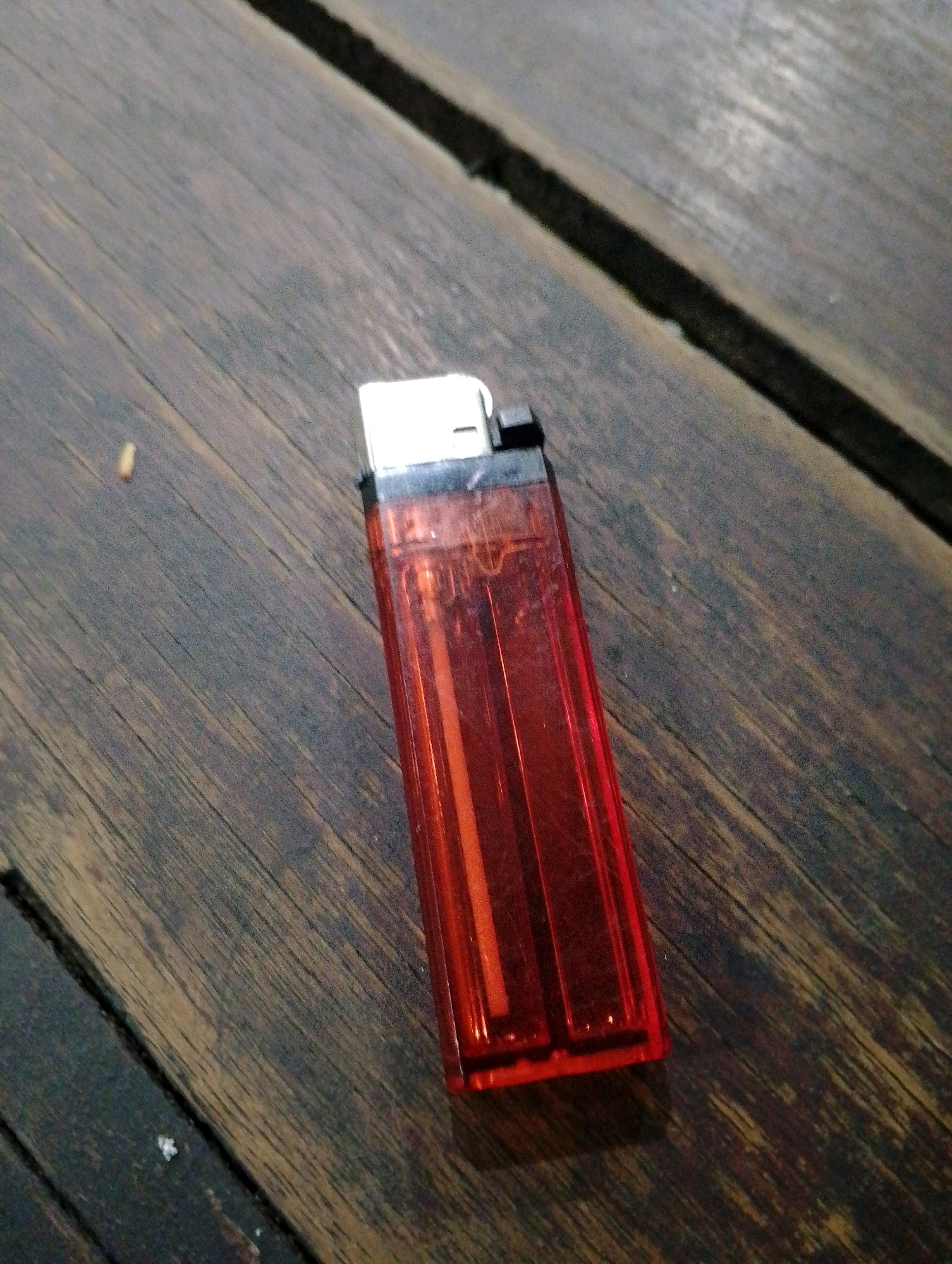 A red disposable lighter is placed on a dark wooden surface. The lighter is oriented diagonally, with its top portion towards the upper left of the image. The wooden surface appears to be made of planks, with visible grain and texture. There are some dark lines suggesting cracks or grooves between the planks. A faint shadow is cast by the lighter. No people or other objects are immediately apparent in the scene. The lighting suggests an indoor setting, possibly at dusk or with low ambient light. The location is Unknown, Unknown.