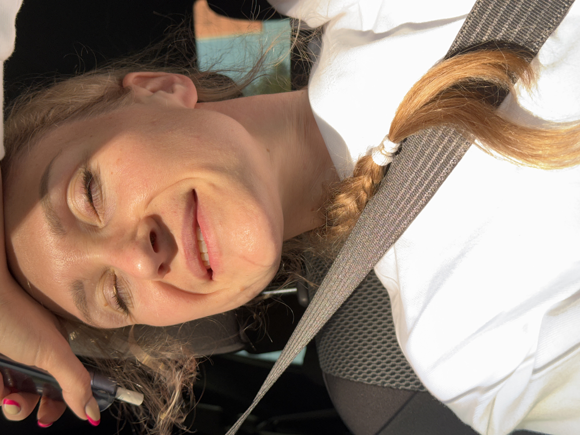 A woman is pictured, seemingly in a car. She wears a white top and a seatbelt is visible across her body. Part of her blonde hair is tied back in a braid. In her hand, she holds a cylindrical object. No information suggests the location is Chaikovsky, Russia.