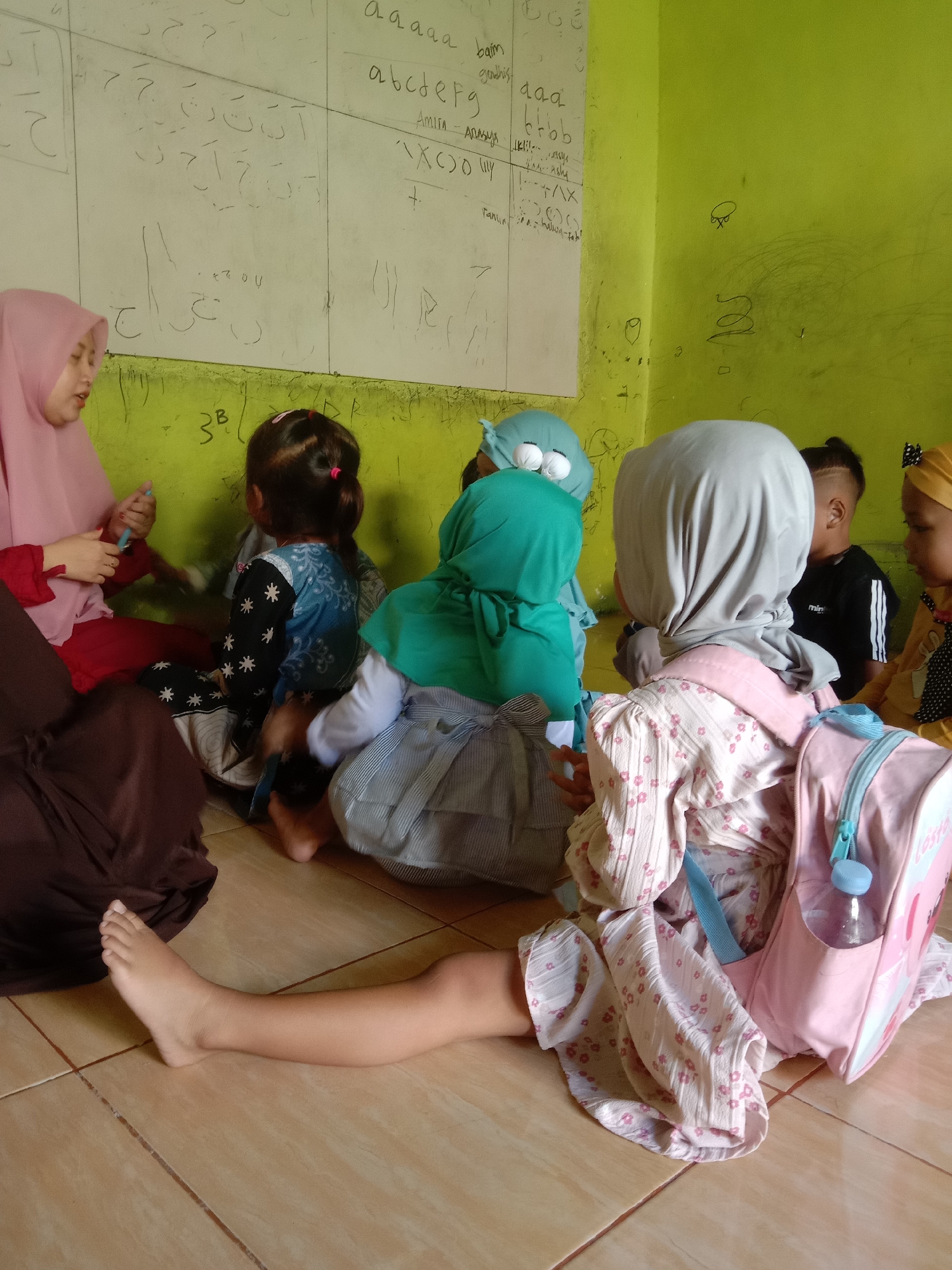 This is a medium shot, eye-level, indoor photograph of a group of young children and a woman sitting on a tiled floor in front of a whiteboard. The room is painted a bright lime green. On the whiteboard, there are various writings, including what appear to be Arabic letters and some English letters like "a a a a", "b c d e f g", and "b b b". Some of the children are wearing headscarves. One child in the foreground, facing away from the camera, is wearing a light pink dress with a floral pattern and a light pink backpack with blue accents. A clear plastic water bottle is visible in the backpack's side pocket. The woman, wearing a pink headscarf and a red top, is on the left side of the frame, seemingly interacting with the children. The children are seated closely together, with some looking towards the whiteboard and others towards the woman. The overall scene suggests an educational setting, possibly a classroom. The photograph was taken in Tulungrejo, Indonesia.