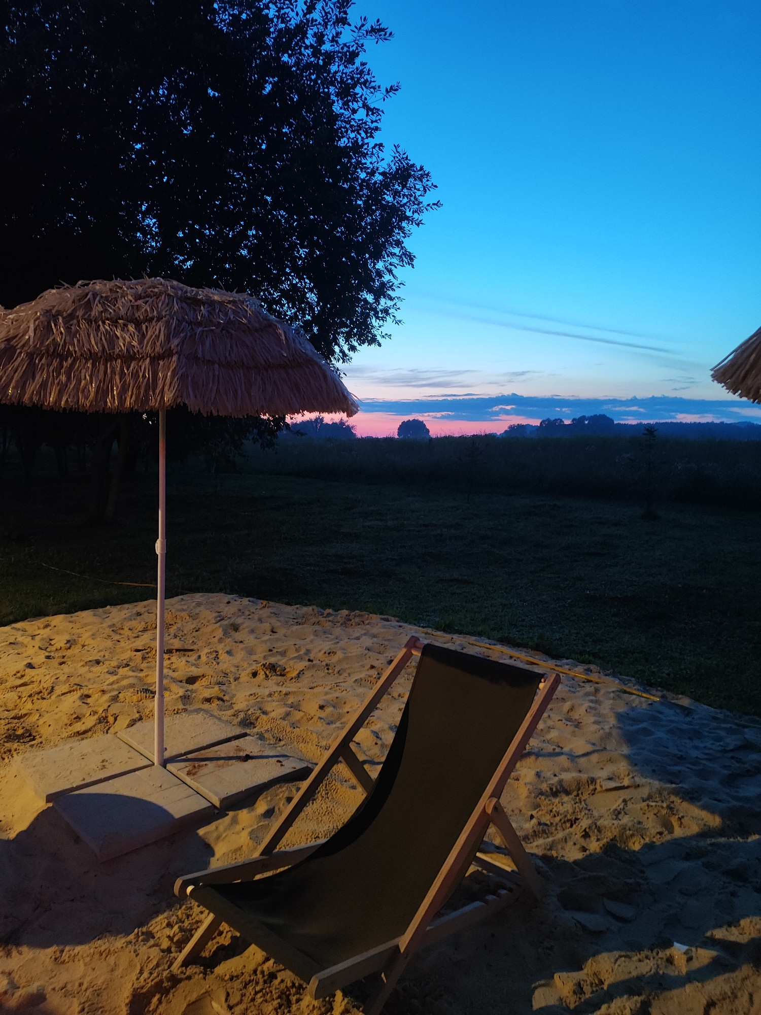 The image depicts a beach chair and a straw umbrella on a sandy area, likely at dusk or dawn. The background shows a grassy field and a tree silhouette against a twilight sky. The sky is a gradient of deep blue at the top, transitioning to lighter shades of blue and pink near the horizon, with wispy clouds. The sandy area appears to be part of a recreational or outdoor seating area. The scene suggests a peaceful and tranquil setting. This scene is located in Bronnitsy, Russia.
