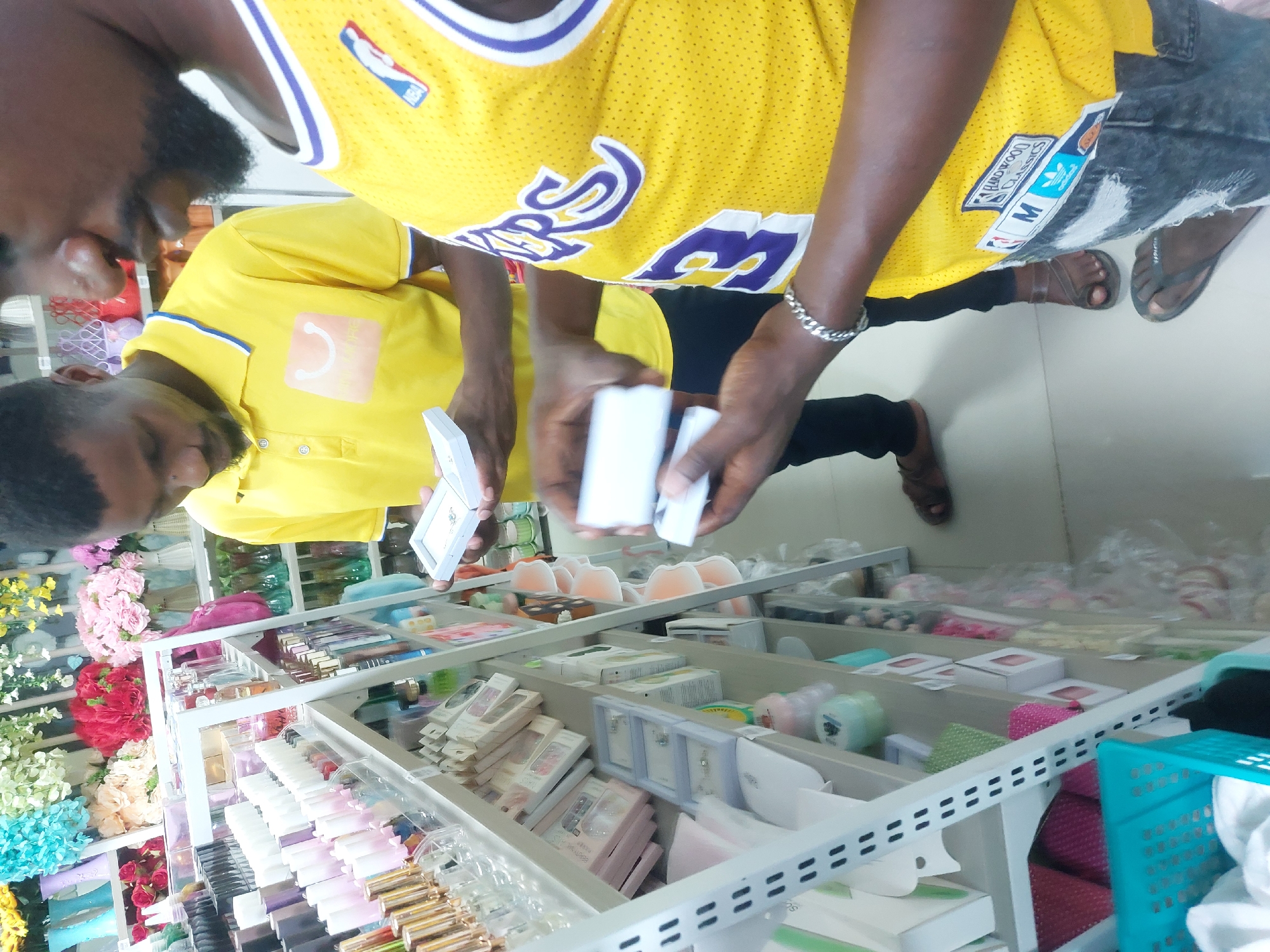 Two African males stand indoors, looking at jewelry. One wears a yellow Lakers jersey and the other a yellow collared shirt. The jewelry is arranged in a white-framed shelf. The location appears to be a retail store. Based on the prompt, it is likely to be Dar es-Salaam, Tanzania.