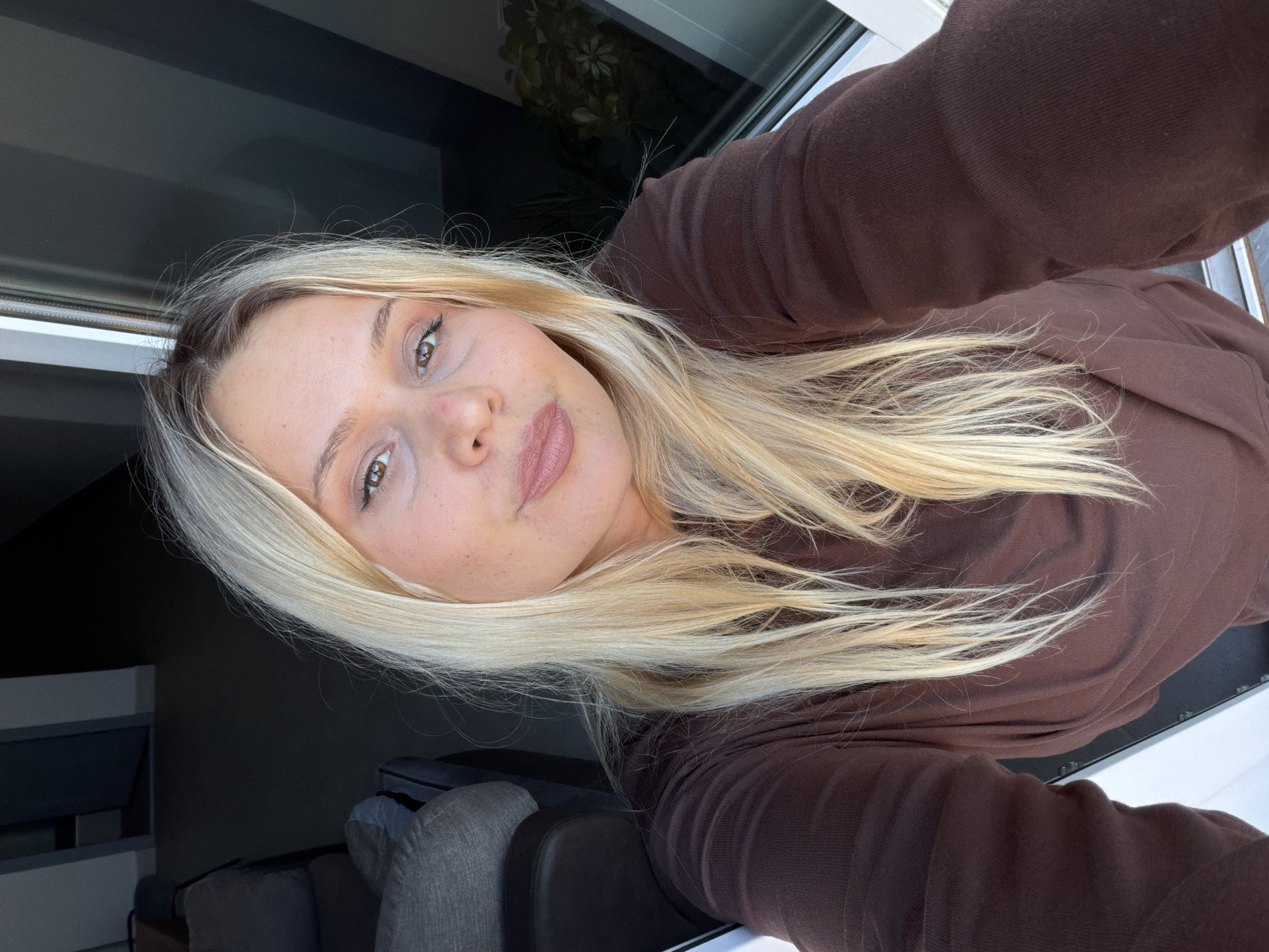 A young woman with blonde hair is taking a selfie indoors. She's wearing a brown long-sleeved shirt and her hair is styled in loose waves. The background shows a window with plants visible outside and a glimpse of a gray sofa. The location in the city of Sarapul, Russia is not discernible from the image.