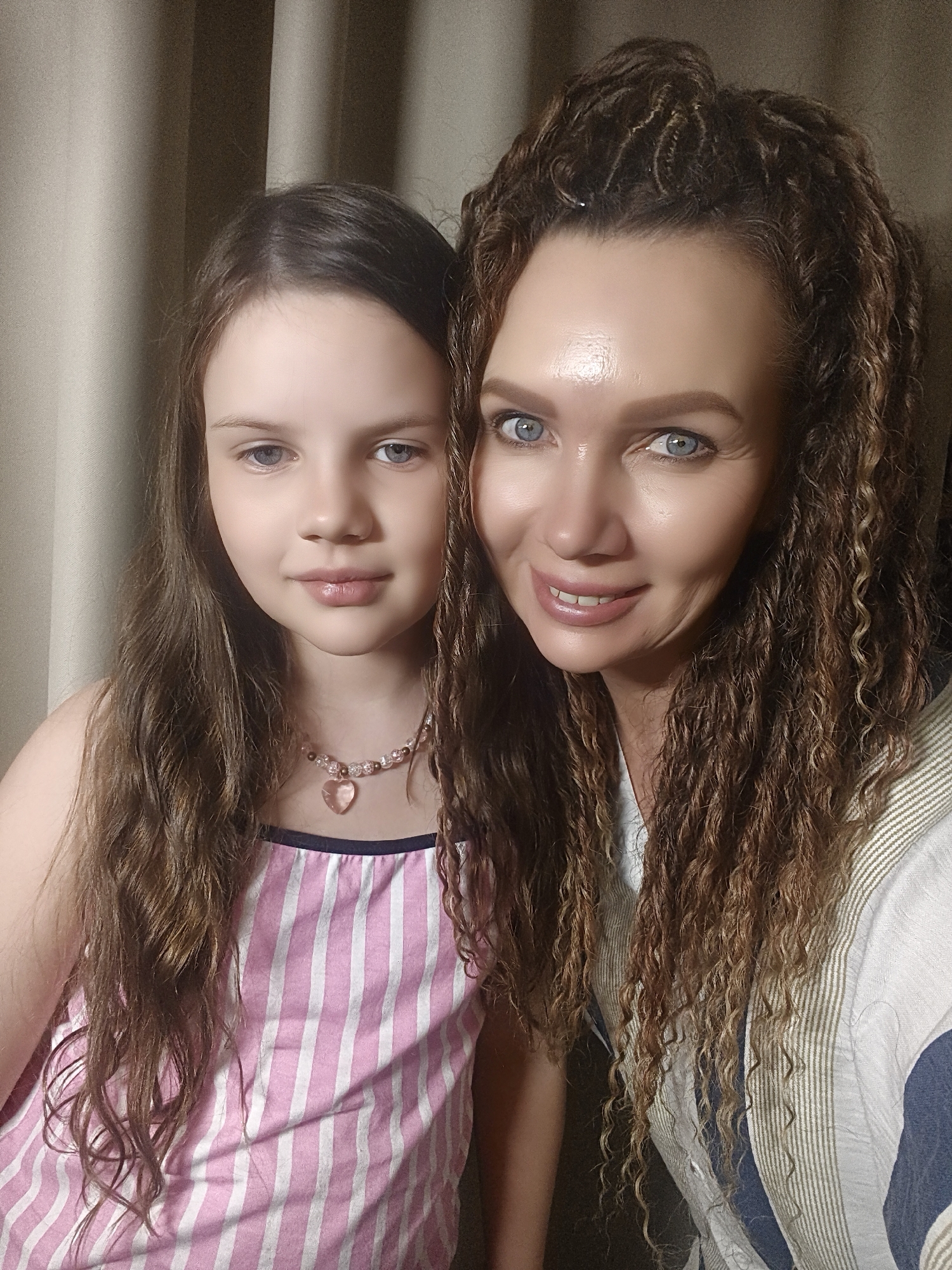 A medium shot depicts two Caucasian females, a child and an adult, posed closely together. The child, wearing a pink and white striped top with a dark blue neckline, has straight, long brown hair and a pink bead necklace with a heart pendant. The adult, likely the child's mother, has dark brown, curly hair. Both have fair skin and light blue eyes. The adult is wearing a cream-colored shirt with striped accents. The background is a plain brown curtain. The setting appears to be indoors. The location is Unknown, Unknown.