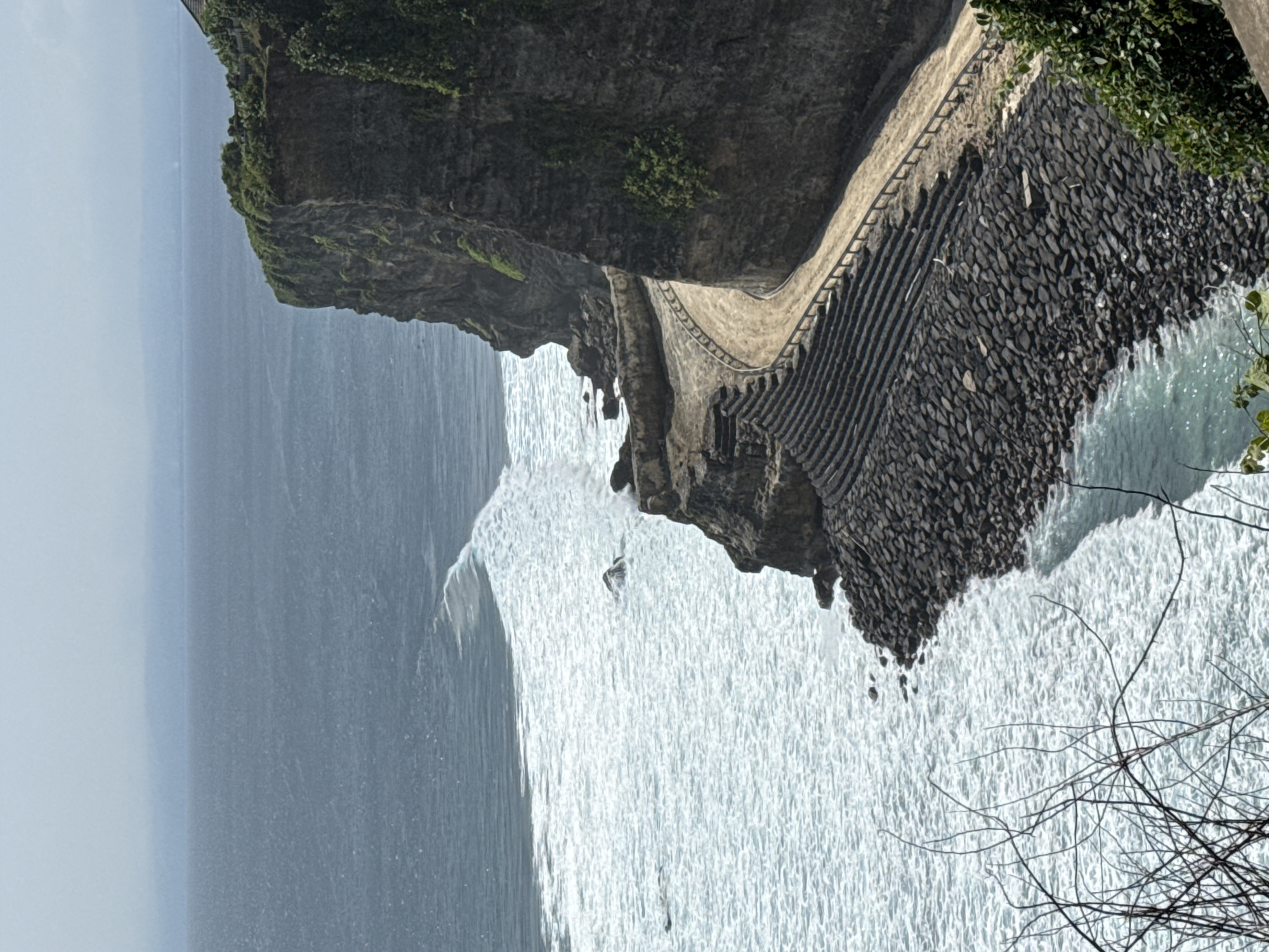The image depicts a rugged coastline in Kuta Selatan, Indonesia. A rocky cliff face, partially covered in vegetation, meets the ocean. A stone walkway with steps is built along the cliff's edge, leading towards the water. The ocean is visible, with waves crashing against the base of the cliff. In the foreground are some bare branches.
