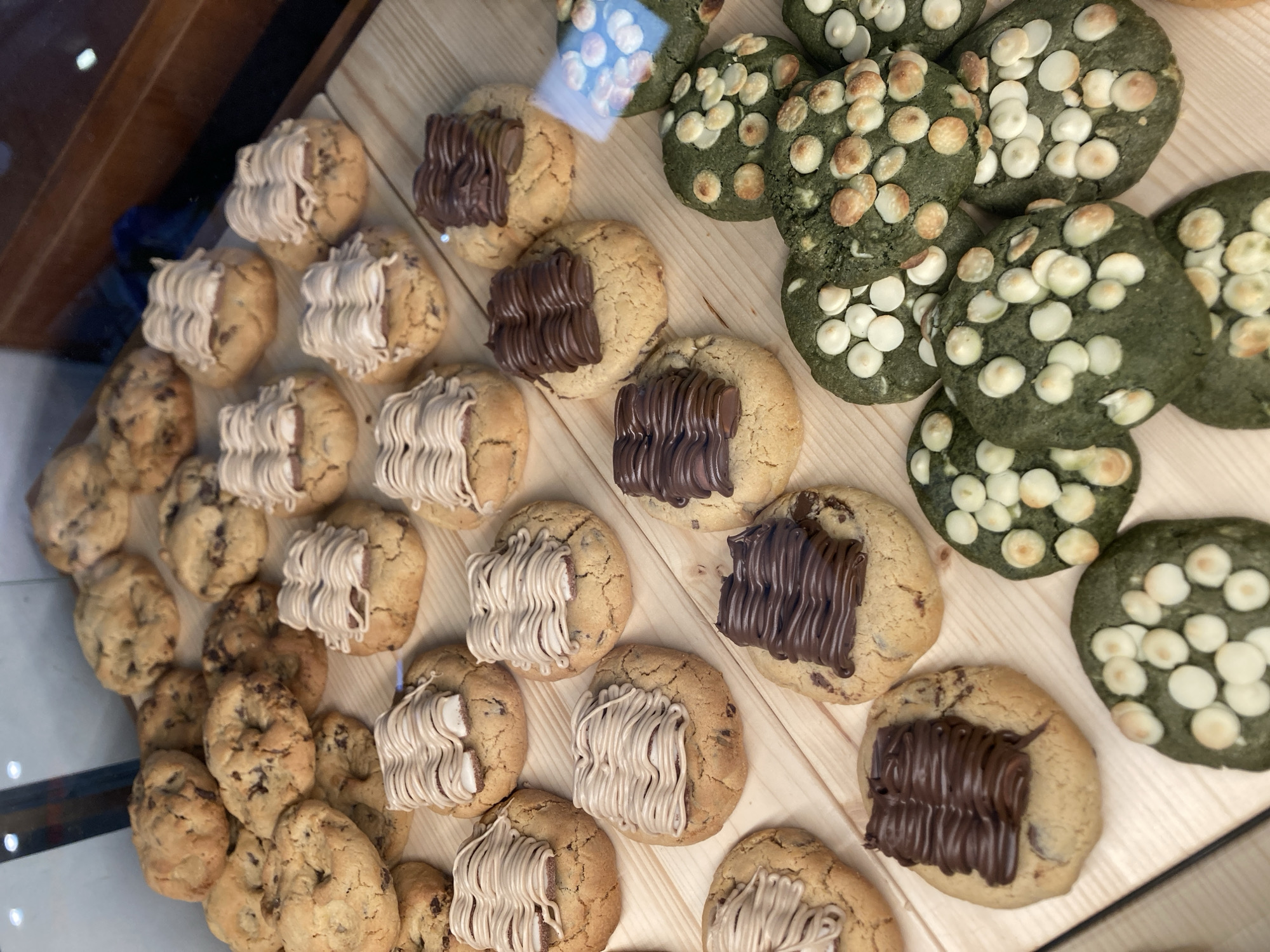 The image displays an assortment of cookies arranged on a wooden surface, likely a display case or counter. The cookies are varied in type. Some are classic chocolate chip cookies. Others are adorned with white frosting drizzled in a wavy pattern. A third type are dark green cookies studded with white chocolate chips. Several cookies are topped with a thick layer of melted chocolate, also drizzled in a wavy pattern. One cookie in the upper left corner has a small, clear plastic bag containing white chips. The setting appears to be a bakery or dessert shop, given the presentation of the baked goods. The lighting suggests an indoor environment, possibly during daytime. No people are visible in the frame, but the arrangement implies a commercial setting. The overall impression is one of a visually appealing selection of sweets. There is no discernible text within the image.