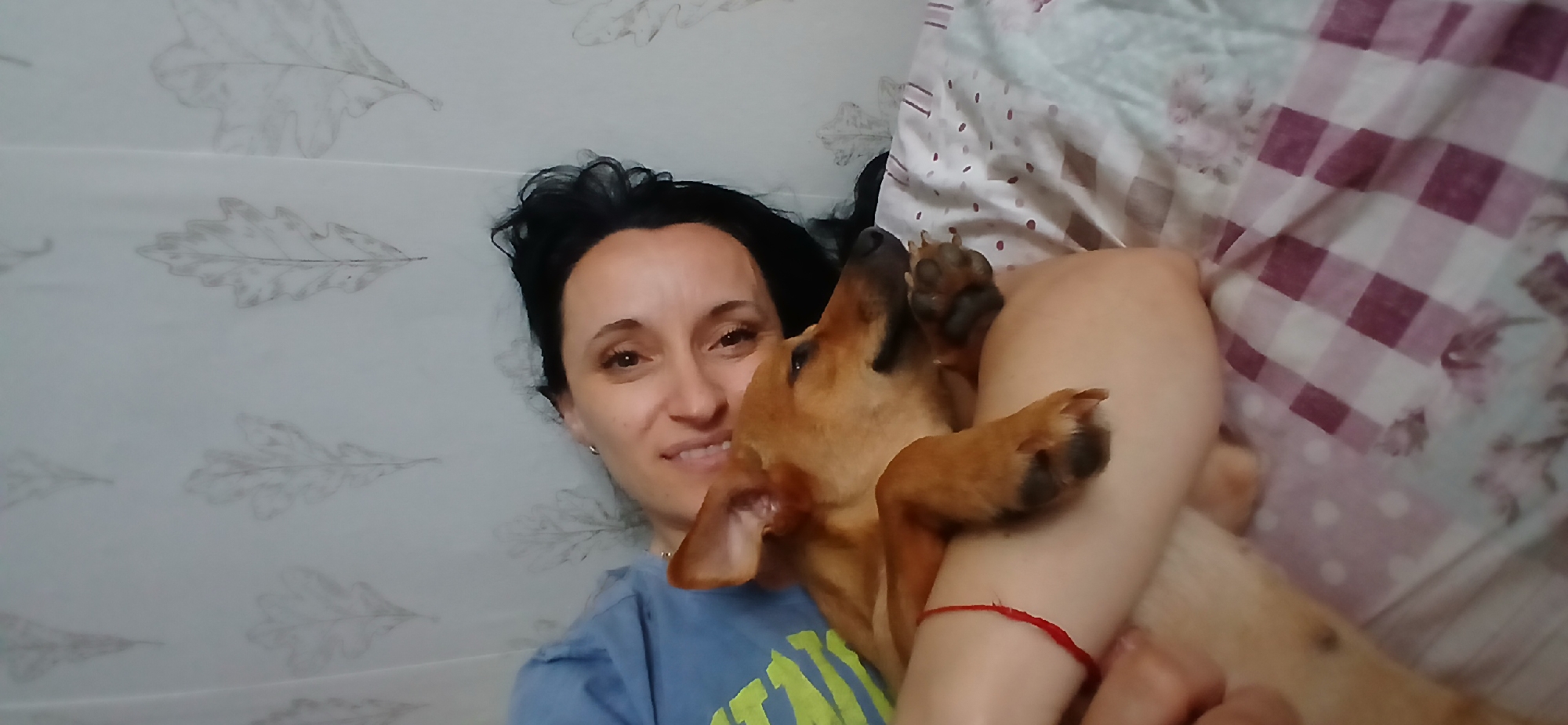 The image depicts a woman and a dog in a relaxed, indoor setting. The woman, with dark hair, is lying down and smiling as she holds a small, light brown dog. The dog is nestled into her arms, with its head resting on her. Both appear comfortable and affectionate. They are on a bed covered with a checkered duvet in shades of purple and white, with a floral pattern visible in some sections. The background shows a wall with a wallpaper featuring a subtle, grey leaf motif. The overall atmosphere is one of intimacy and comfort. The image does not provide any specific information to indicate its location in Blagoevgrad, Bulgaria, or any other city.