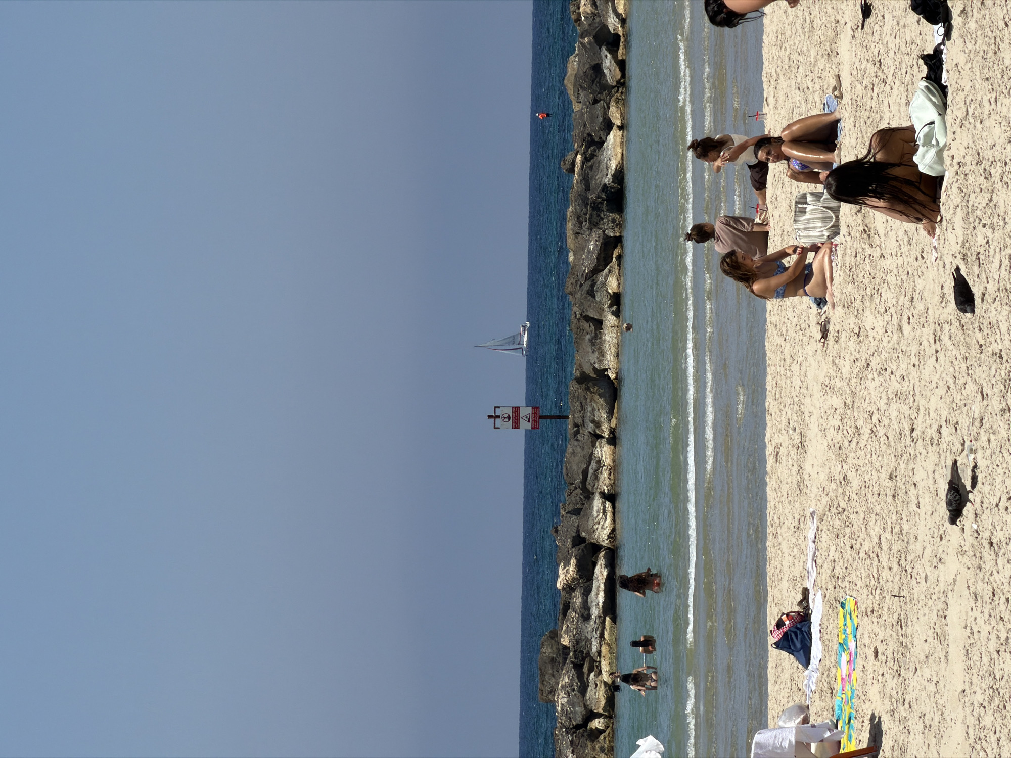A wide shot captures a sunny day at a Tel Aviv beach with a clear blue sky and calm water. The scene is divided into the beach and the sea, separated by a rocky breakwater.
On the sandy beach, several women are relaxing or sunbathing. In the foreground, a woman with long dark hair sits facing away from the camera, with a light green backpack and a dark bag beside her. Further back, a group of women are seated on towels. One woman in a blue bikini adjusts her foot, another sits with her legs extended, and two others sit facing away from the camera. A blue and white striped bag sits between two of the women. A pigeon is visible on the sand in the lower left quadrant of the image. Scattered items like towels and bags are present on the beach.
In the sea, several women are wading or swimming. In the mid-ground, three women are in the water, with one in the foreground and two further back. Further out in the sea, a sailboat is visible on the horizon to the left, and a person in an orange life vest is visible to the right. The rocky breakwater runs horizontally across the middle of the image, with a red and white sign on a pole situated on it. The water is a clear, turquoise color, with gentle waves lapping at the shore. The overall impression is a relaxed day at the beach.