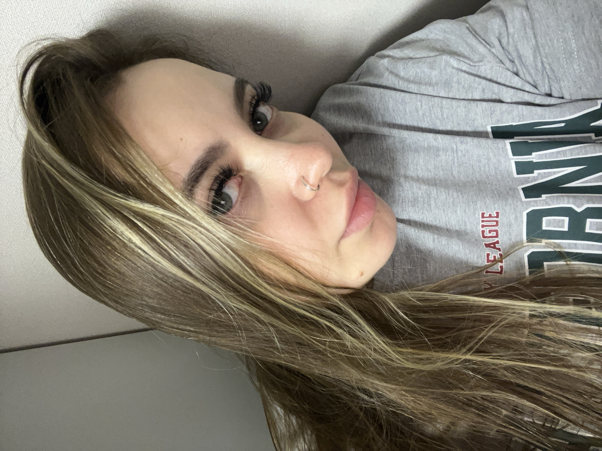 A young woman with long, blonde-highlighted brown hair is lying down, facing the camera. She has a nose ring and visible eyelashes. She is wearing a gray t-shirt with dark green and red lettering. The background appears to be a plain wall. There is no indication of a location in Krasnodar, Russia.