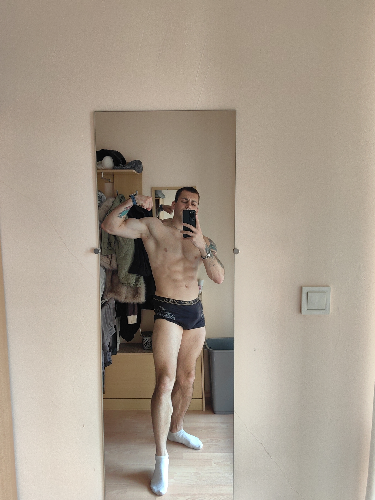 A Caucasian male is standing in front of a full-length mirror, taking a selfie with his phone. He is wearing dark blue boxer briefs and white socks. He is flexing his right bicep, with his arm bent at the elbow and his fist clenched. His left arm is bent at the elbow and his hand is resting on his hip. He has visible abdominal muscles and tattoos on his right arm. The mirror is reflecting the interior of a room, which appears to be a bedroom or dressing area. In the background, there is a closet with clothes hanging and shelves with various items. There is also a wooden dresser and a gray trash can visible. The walls are painted a light peach color, and the floor is wooden. A light switch is visible on the right wall. The scene is consistent with an indoor residential setting, potentially in Varna, Bulgaria, given the prompt's location specification.