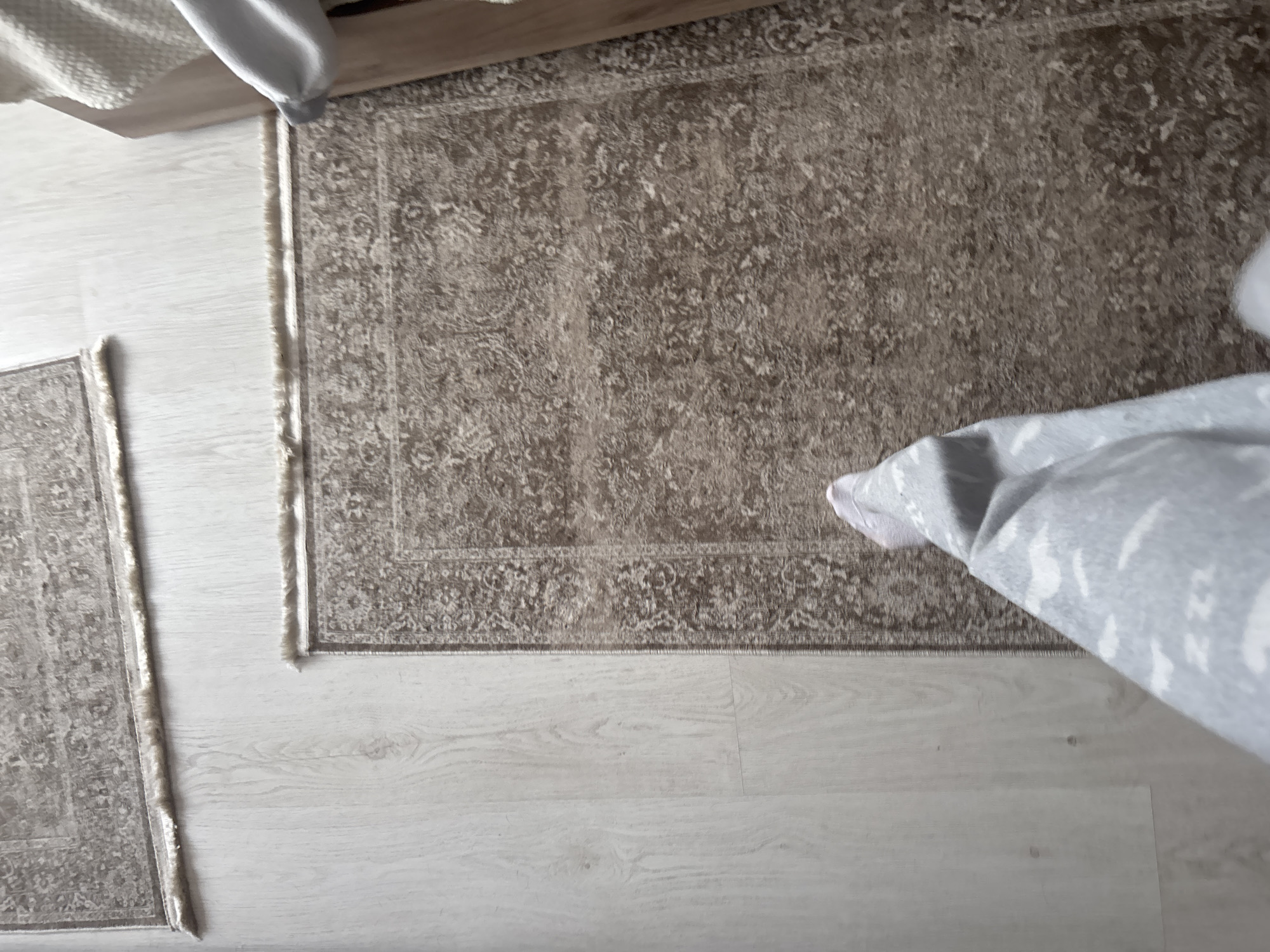 The image shows a close-up, high-angle view of two rugs on a light-colored wooden floor. The rug in the foreground is a medium-sized, patterned rug with a distressed, faded design in shades of brown and beige. Its edges have a fringed trim. A person's lower legs and feet, clad in grey patterned pajamas and white socks, are visible in the bottom left corner of the image, suggesting the person is standing or walking on the rug. Another, larger rug with a similar patterned design is partially visible in the upper portion of the image. To the right, a light-colored wooden furniture piece, possibly the side of a bed frame or sofa, is visible. The lighting suggests an indoor setting, likely a home environment. The location is specified as Unknown, Russia.