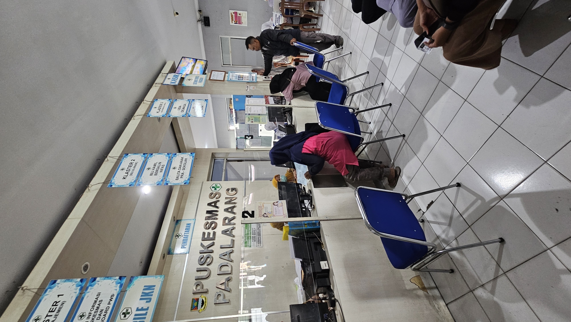 The image depicts the interior of a public health center, likely a Puskesmas, in Kertamulya, Indonesia. The scene is a waiting area with several people. In the foreground, blue chairs with metal frames are arranged on a tiled floor. The walls are painted in a light color, and there are signs hanging from the ceiling indicating different service clusters and registration areas. A prominent sign reads "PUSKESMAS PADALARANG."
Several individuals are visible. In the center, a person wearing a dark hijab and a pink shirt is seated at a counter, appearing to interact with a staff member behind a glass partition. Another staff member, wearing a yellow hijab, is also visible behind the counter. To the right, a man in a dark jacket is standing and gesturing, seemingly addressing someone. Other individuals are seated or standing further back, suggesting a busy service day. The overall environment appears to be functional and designed for public service, with elements like computer monitors and filing systems present at the counters.