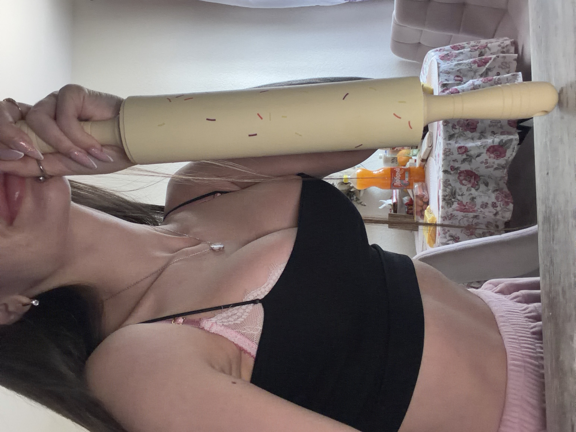 A horizontal indoor shot depicts a person holding a rolling pin with both hands. The rolling pin is beige and decorated with small, multi-colored dashes. The person has fair skin, dark hair, and wears a black top over a pink undergarment. They have a silver necklace and earrings, and their nails are long and manicured. The background includes a table covered with a floral-patterned cloth, an orange bottle labeled "Добрый" (likely Russian), and furniture with pink accents. The location cannot be determined from this image.
