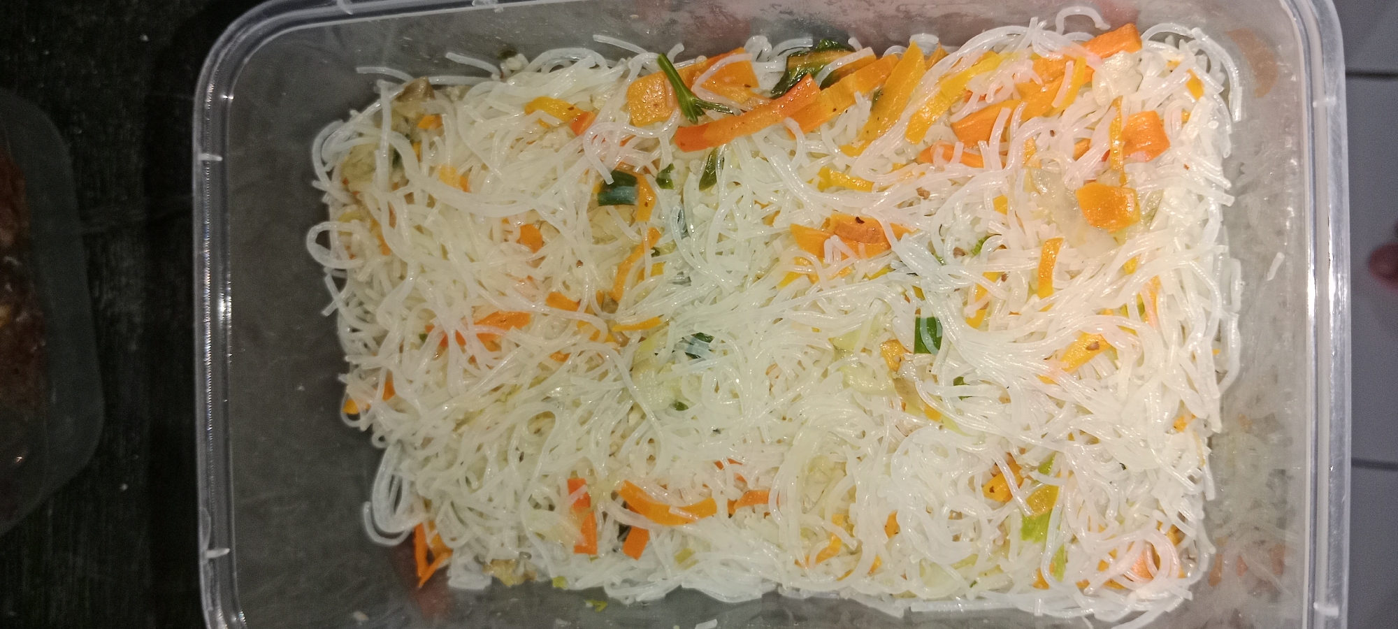 A clear plastic rectangular food container holds a portion of white rice vermicelli mixed with julienned carrots and green onions. The food is viewed from directly above. The container is placed on a dark, textured surface that appears to be a countertop. To the left of the container, a dark blurry object is partially visible, which could be another container. The background is out of focus and appears to be a domestic or commercial kitchen setting in Subang, Indonesia.
