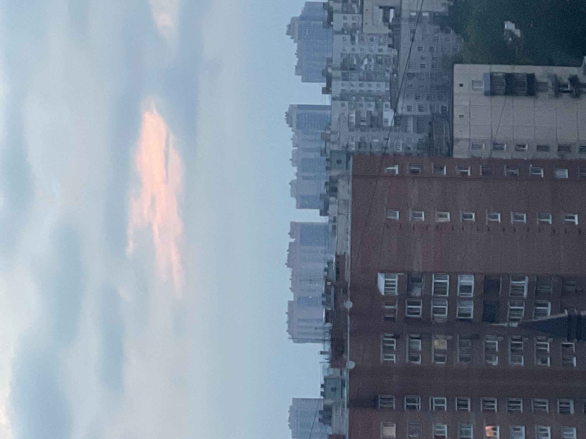 The image shows a cityscape with several apartment buildings under a cloudy sky. In the foreground, a tall brick apartment building dominates the left side of the frame, displaying numerous windows arranged in a grid pattern. To the right of the brick building, other apartment buildings of varying heights and colors are visible, with some appearing older and made of concrete. In the background, a row of much taller, lighter-colored apartment buildings stretches across the horizon, some partially obscured by the sky. The sky itself is overcast with soft, diffused light, and a patch of pinkish-orange color suggests either the sun setting or rising behind the clouds. The overall impression is of a dense urban environment, possibly in the evening or early morning.