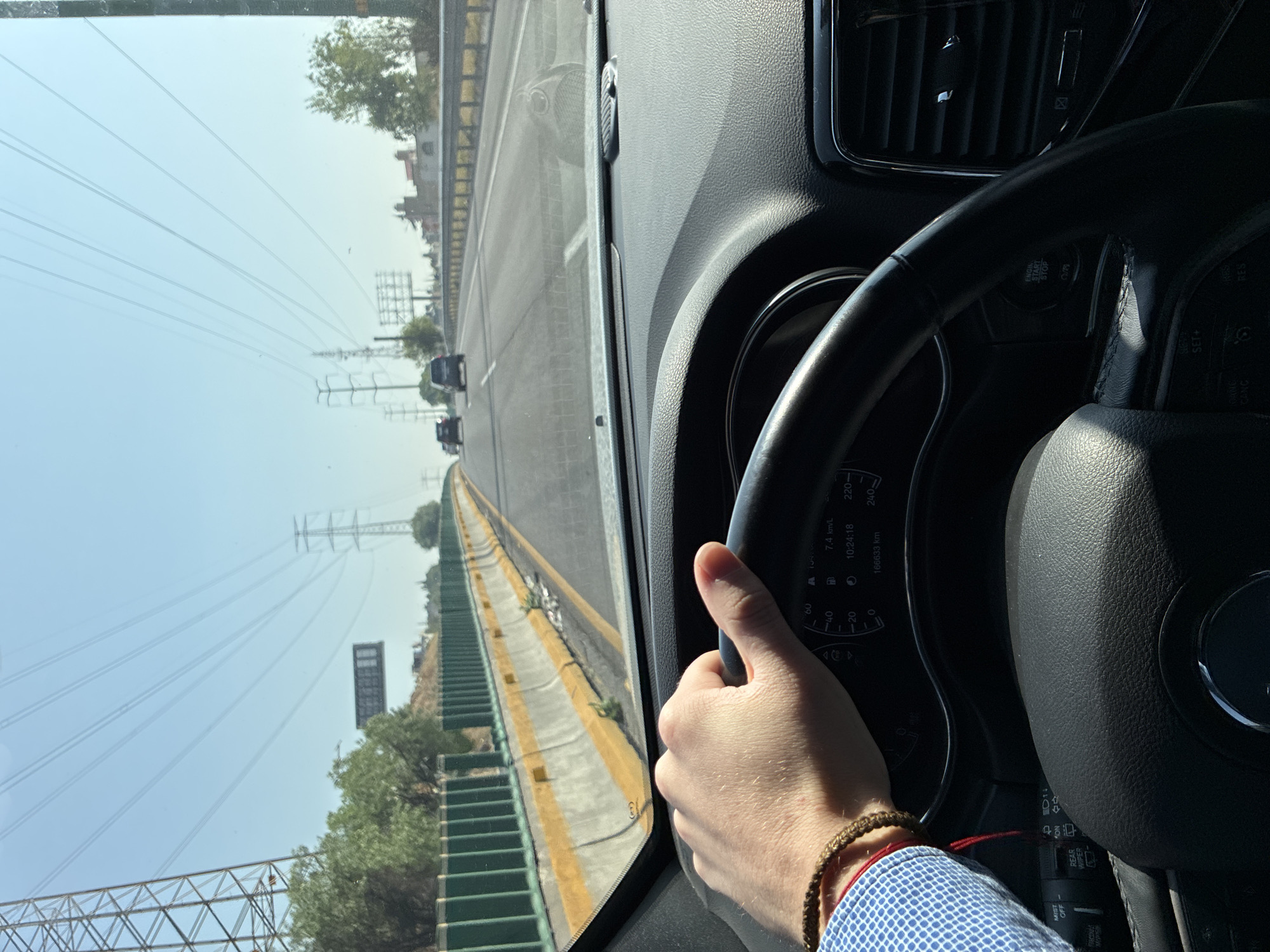 A person is driving a car on a highway, looking out the front windshield, driving on a highway next to a concrete divider wall. There is green foliage on the other side of the wall. Power lines and electrical towers are visible in the distance. The location is Ciudad López Mateos, Mexico. The car's dashboard and steering wheel are visible. The driver's hand is visible, holding the steering wheel, and the driver is wearing a light blue shirt. The car's dashboard displays a time of 10:24:18 and a mileage of 166633 km.
