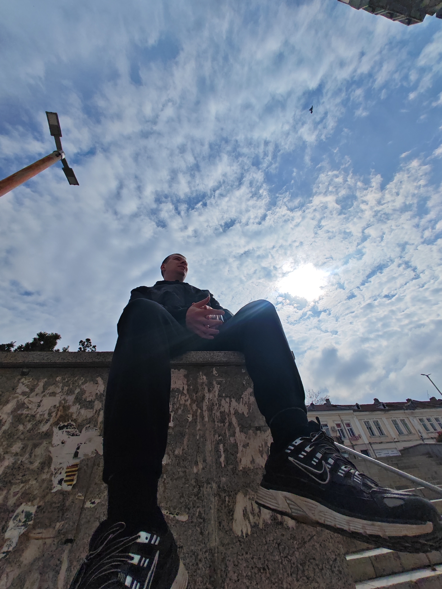A low-angle urban scene in Shumen, Bulgaria, captures a male individual sitting atop a weathered concrete retaining wall. The wall surface is marred by remnants of torn paper and posters. The subject is dressed in black athletic attire, comprising a long-sleeved top and trousers, and wears black Nike athletic shoes with prominent white swoosh logos and white/light grey soles. His hands are clasped, and his gaze is directed upward towards the sky, where the sun is visible through scattered white clouds, creating a bright flare. A small, dark bird is silhouetted in flight against the blue and white sky. To the left of the frame, a rust-colored pole with two horizontal, rectangular light fixtures extends upwards. In the background on the right, traditional multi-story buildings with light-colored facades and red tiled roofs are visible, alongside a grey metal handrail and stone steps descending from the elevated position of the wall. Dark green coniferous trees are also present behind the individual.