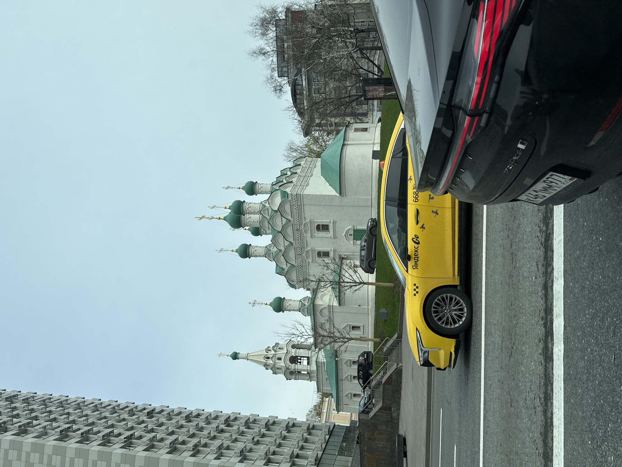 The image, taken from within a vehicle, captures an urban street scene in Moscow, Russia, featuring a juxtaposition of historic and modern architecture alongside contemporary vehicles.
In the immediate foreground, partially obscuring the view, is the black rear bumper and taillight of a car, which appears to be an Exeed model with the license plate "K484MM977 RUS". This plate indicates registration in Moscow. Adjacent to this car, and more prominently featured, is a yellow Yandex Go taxi. The taxi displays "Яндекс Go" and the number "668" on its side. Both vehicles are situated on an asphalt road with white lane markings.
The central and most striking element of the scene is a magnificent white Russian Orthodox church with multiple green, onion-shaped domes, each topped with a golden cross. The church exhibits intricate architectural details. Several bare-branched trees are scattered around the church, suggesting either late autumn, winter, or early spring.
To the left of the frame, a tall, modern building with a distinctive, patterned grey facade rises, contrasting sharply with the traditional church architecture. The sky above is uniformly overcast and light grey, indicating a cloudy day.