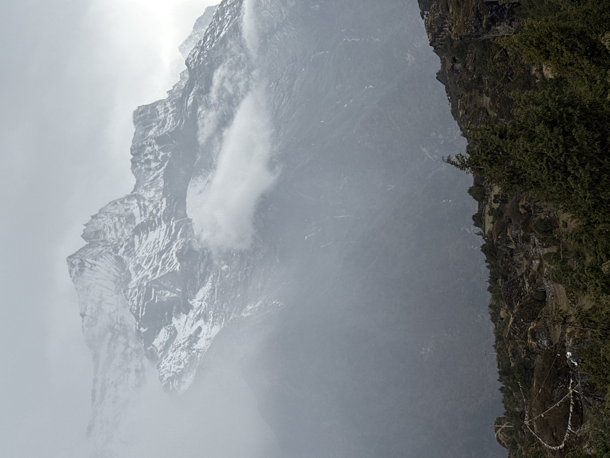 The image depicts a mountainous landscape in what is likely the region of Namche, Nepal. Dominating the left side of the frame is a massive, snow-capped mountain obscured by heavy fog and clouds. To the right, a steep, rocky cliff covered in vegetation descends. In the foreground, prayer flags are visible, hanging from the cliff's edge. The overall atmosphere is one of overcast and potentially cold conditions.
