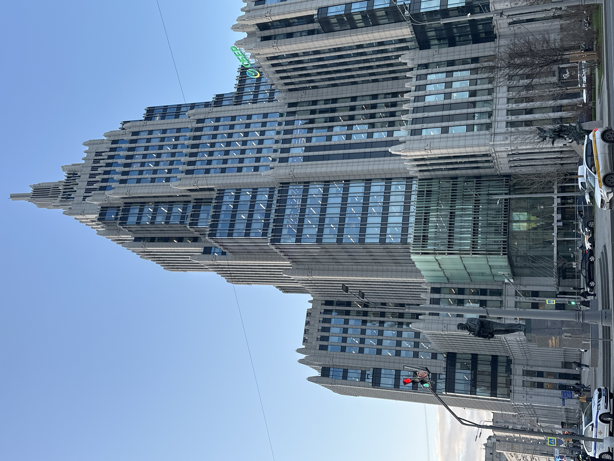 The image depicts a modern skyscraper in Moscow, Russia, with a distinctive stepped facade of glass and light-colored stone or concrete. A green and blue sign is visible on the upper portion of the building. A bronze statue stands on a pedestal in front of the building, adjacent to a city street. Cars are parked along the street, including what appears to be a white police car. Traffic lights are present, with one showing a red light. The sky is clear and blue. Pedestrians are visible on the sidewalks, indicating an urban setting.