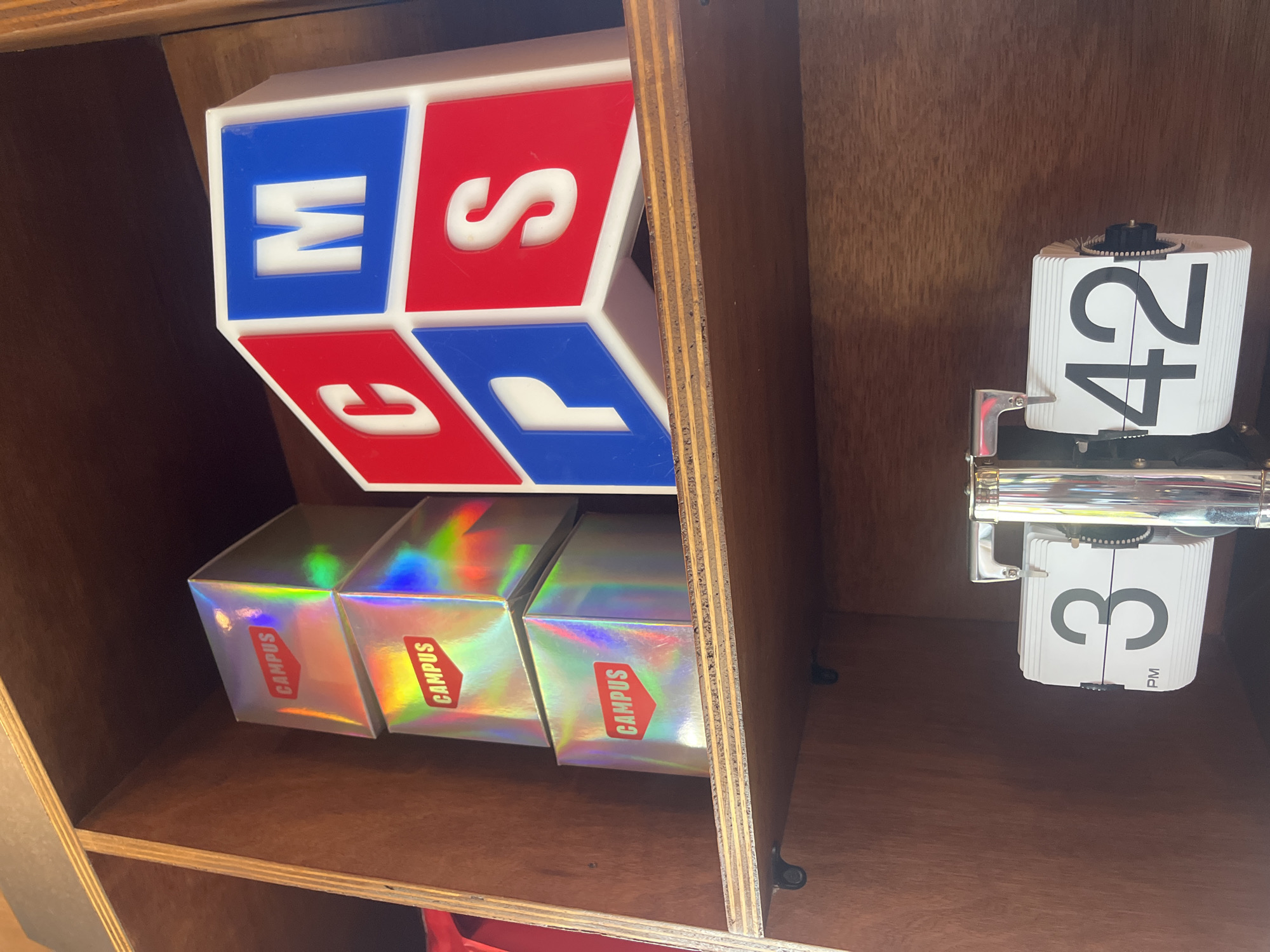 Inside a wooden cabinet, a scene unfolds with distinct elements. On the left, four cubes catch the eye. A larger cube, adorned with blue and red blocks displaying the letters "W," "S," "C," and "P," sits above three smaller, silver-holographic boxes labeled "CAMPUS." To the right of the cubes is a unique, white flip clock displaying "42" on the top part and "3 PM" on the bottom part. The setting is North Kuta, Indonesia. No people are visible in the frame, with the focus solely on these inanimate objects within the cabinet.