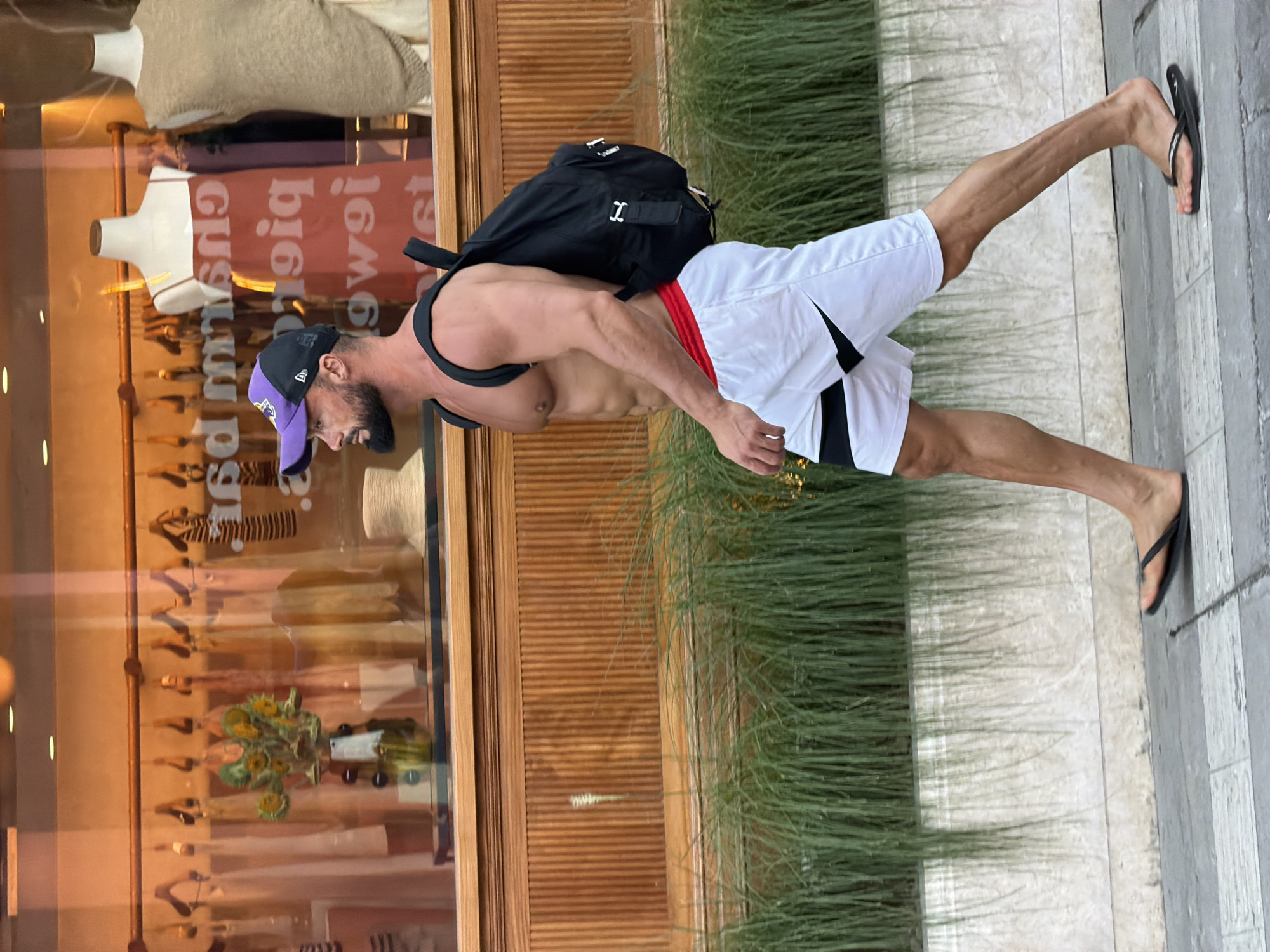 An athletic man with a beard, wearing a purple and black baseball cap, a black backpack, red athletic briefs visible above white athletic shorts with black accents, and black flip-flops, walks from left to right on a sidewalk. He is shirtless, revealing a muscular physique. Behind him, a storefront window displays clothing on mannequins and a floral arrangement. A row of tall, green plants borders the sidewalk in front of the storefront. The scene appears to be set in a city, but the specific location of Mengwi, Indonesia, is not evident from the visual information in the image.