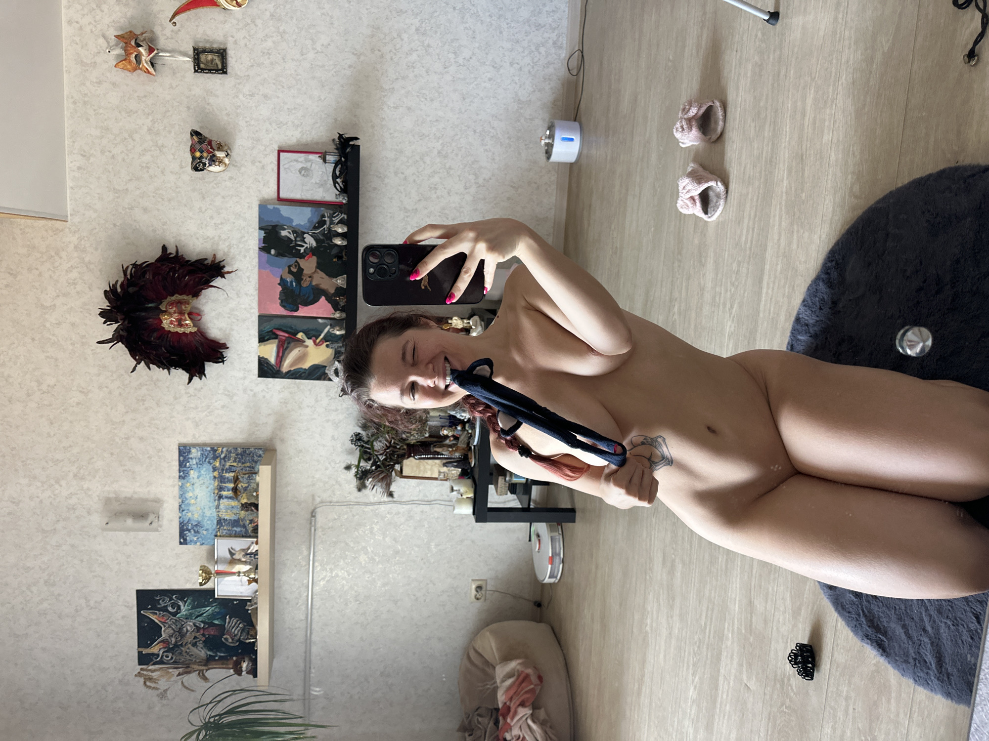 A fair-skinned woman, nude and smiling, takes a selfie in what appears to be a living room in Vladimir, Russia. She holds a black smartphone with a dark case in her right hand, positioned above her chest, capturing her upper body from a high angle. Her hair is tied back, with some pinkish-red strands visible. Her left arm is raised, with a blue or black cloth object in her left hand and tucked into her mouth. A tattoo is visible on her left arm/shoulder area.
The background shows a cluttered room with light-colored wallpaper and wooden flooring. On the wall, there are various decorative items including several Venetian-style masks, two small paintings or prints, and a framed picture. Below these, a shelf holds more decorative objects and small figurines. To the right of the woman, on the floor, are two light pink slippers and a dark grey or black circular rug. A white cylindrical device with blue light, possibly an air freshener or diffuser, is on the floor near the wall. In the lower left corner, a cream-colored beanbag chair is visible with some clothes or fabric on it. A white robotic vacuum cleaner is visible further back in the room, near a wall outlet. The overall impression is an indoor, casual setting.