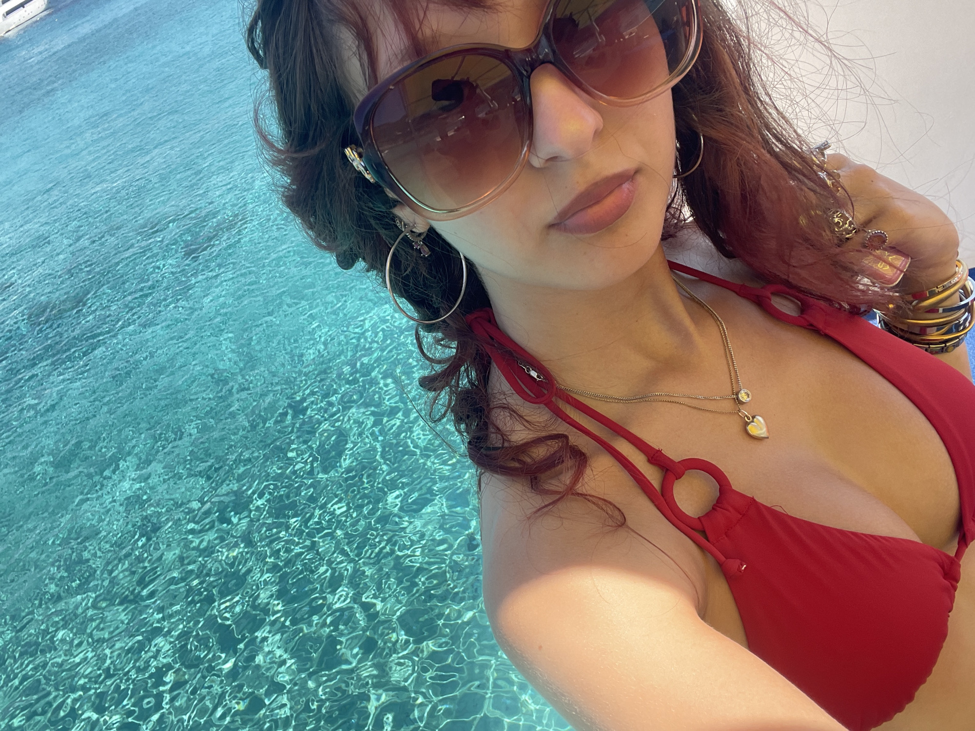 A woman, viewed from the chest up, poses for a selfie by a pool. She is wearing a red bikini top, large sunglasses, and jewelry. Her hair is dark brown with reddish highlights and is slightly damp. The water in the background is clear and blue, with sunlight glinting off the surface. The scene suggests a vacation or leisure setting, possibly in a location like Neo Chorio, Cyprus, known for its coastal beauty and swimming pools.