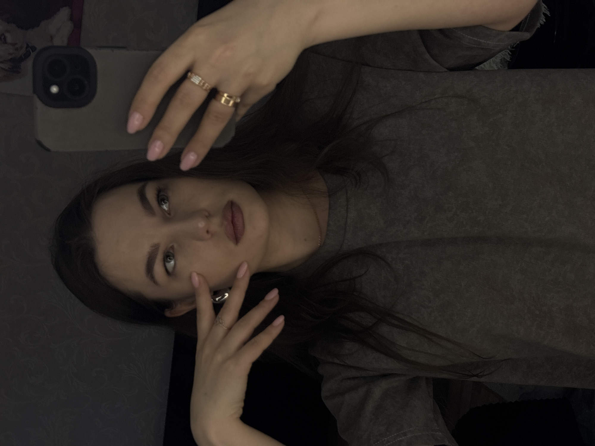 A young woman is taking a selfie. She is wearing a gray T-shirt and has long, dark hair. She is holding a smartphone in her left hand, and her right hand is near her face. She is wearing multiple rings on both hands. The background is dark and indistinct. It is not possible to determine the location as Chaikovsky, Russia, from this image.