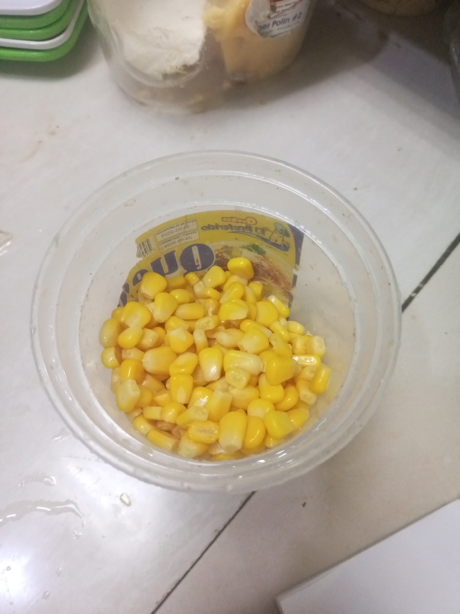 A plastic container filled with cooked corn kernels is centered on a white surface. A yellow package is visible behind the corn inside the container. Other containers are visible in the background. The image appears to be taken indoors in a domestic setting. There is no explicit information linking this image to Juan Griego, Venezuela.