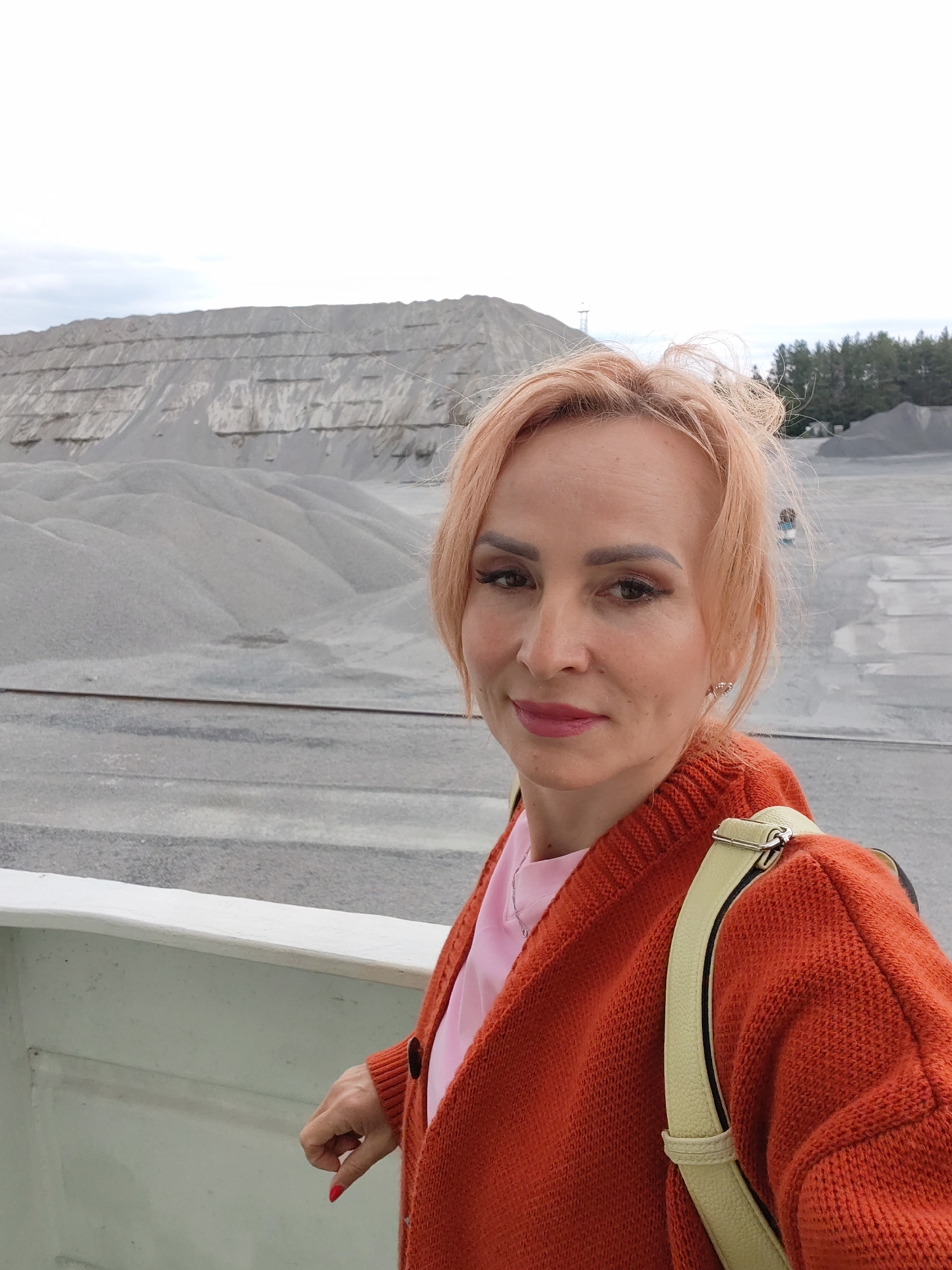 A woman with reddish-blonde hair and makeup, wearing an orange cardigan over a pink shirt, stands on a raised platform. She has a light-colored strap of a bag visible on her shoulder. In the background, there are large mounds of gravel and sand, suggesting an industrial or construction site. The sky is overcast. The overall scene appears to be an outdoor quarry or material storage area. It is not possible to determine the specific location within Shalsky, Russia from the image alone.
