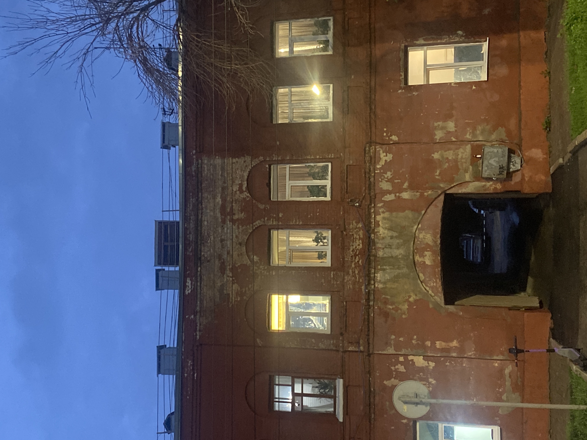 The image shows the facade of a red brick building at dusk. The building has multiple windows, some of which are lit from within, suggesting occupancy. An arched entryway is visible in the center of the building, leading into a dark passageway. To the left of the entryway, a signpost and a scooter are present. The sky above is a deep blue, characteristic of twilight, with a bare tree branch visible on the right side of the frame. The architecture suggests an older building, possibly residential or mixed-use. The overall scene depicts a quiet urban environment in Novokuybyshevsk, Russia, as evening sets in.