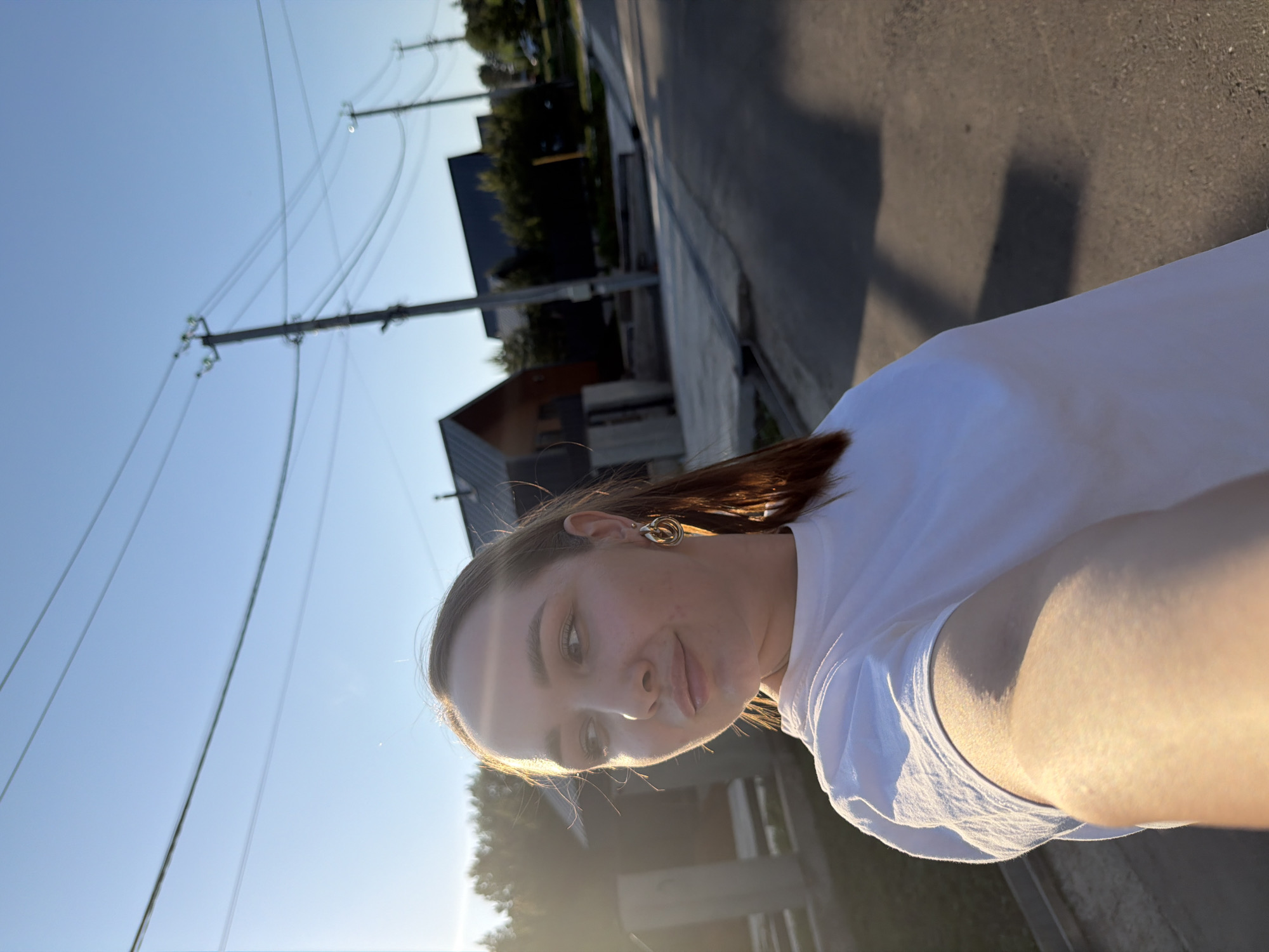 A woman is shown in a portrait setting outdoors with a cityscape visible in the background, possibly in Kamyennoye, Russia. She is wearing a white shirt and a gold earring is visible on her left ear. The setting suggests she is on a street next to a sidewalk with utility poles and buildings in the background. The time of day appears to be early evening or late afternoon, given the presence of low sunlight and shadows. The image is slightly rotated clockwise.