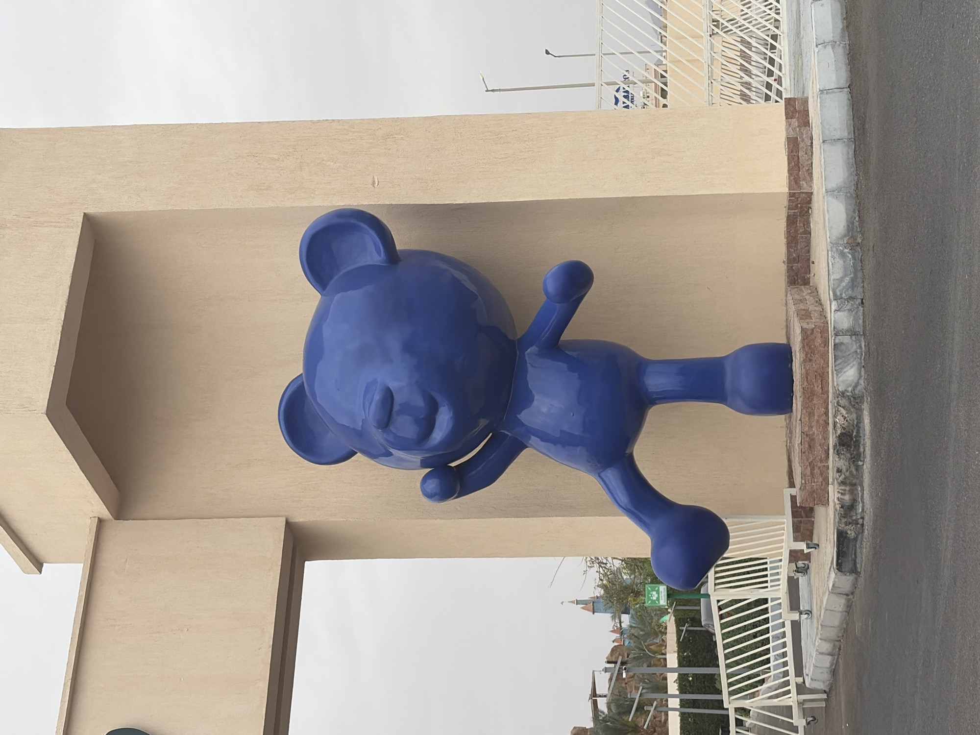 The image shows a large, blue, stylized bear sculpture. The sculpture is positioned at the entrance to a location, seemingly above an archway or entrance feature made of beige material. The bear has a rounded head, sleepy eyes, and a spherical nose. Its arms and legs are stretched out. In the background, partially visible through the archway, is a view of a location in Sharm El Sheikh, Egypt, including palm trees and a castle. A road and a white metal fence are visible. The sky is overcast.