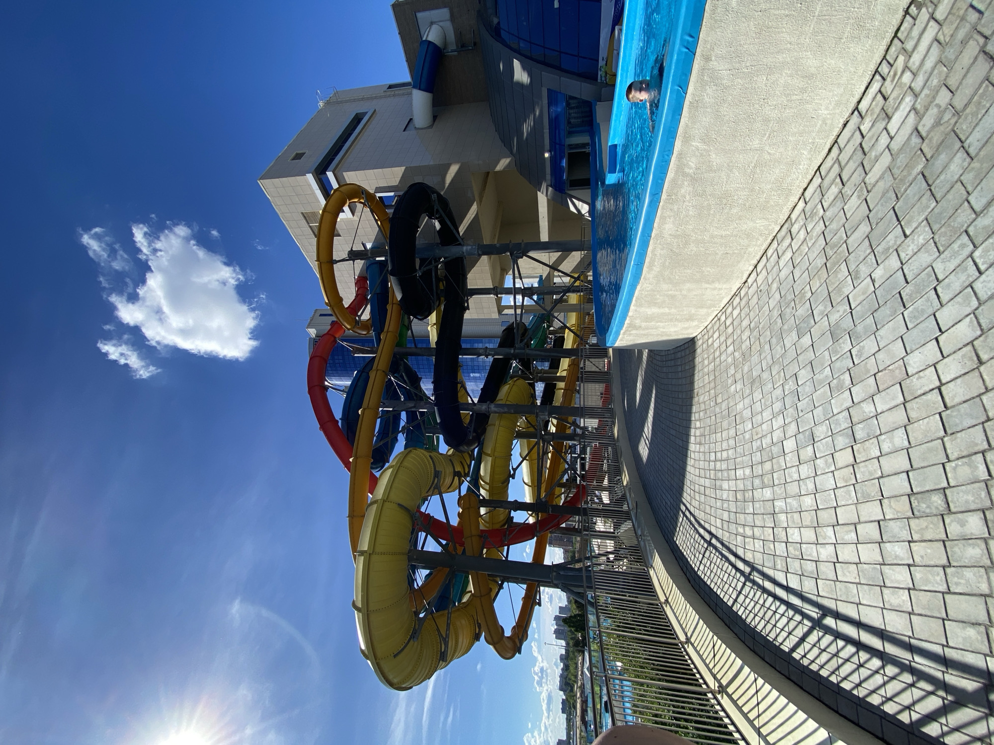 The image captures an outdoor water park scene in Kazan, Russia, under a clear blue sky with a few scattered clouds. The central focus is a complex structure of multicolored water slides, predominantly yellow, red, blue, and black, interconnected and supported by a metal framework. The slides appear to be looping and twisting, suggesting an exciting ride experience. The slides emanate from a light-colored building that is likely the water park's main structure. The person is swimming in a pool near the entrance of one of the waterslides. The foreground features a pathway constructed of light gray paving stones, bordered by a metal railing. In the background, some greenery and the cityscape of Kazan can be seen, hinting at the park's urban location. Sunlight is visible at the bottom of the image, enhancing the brightness of the scene.