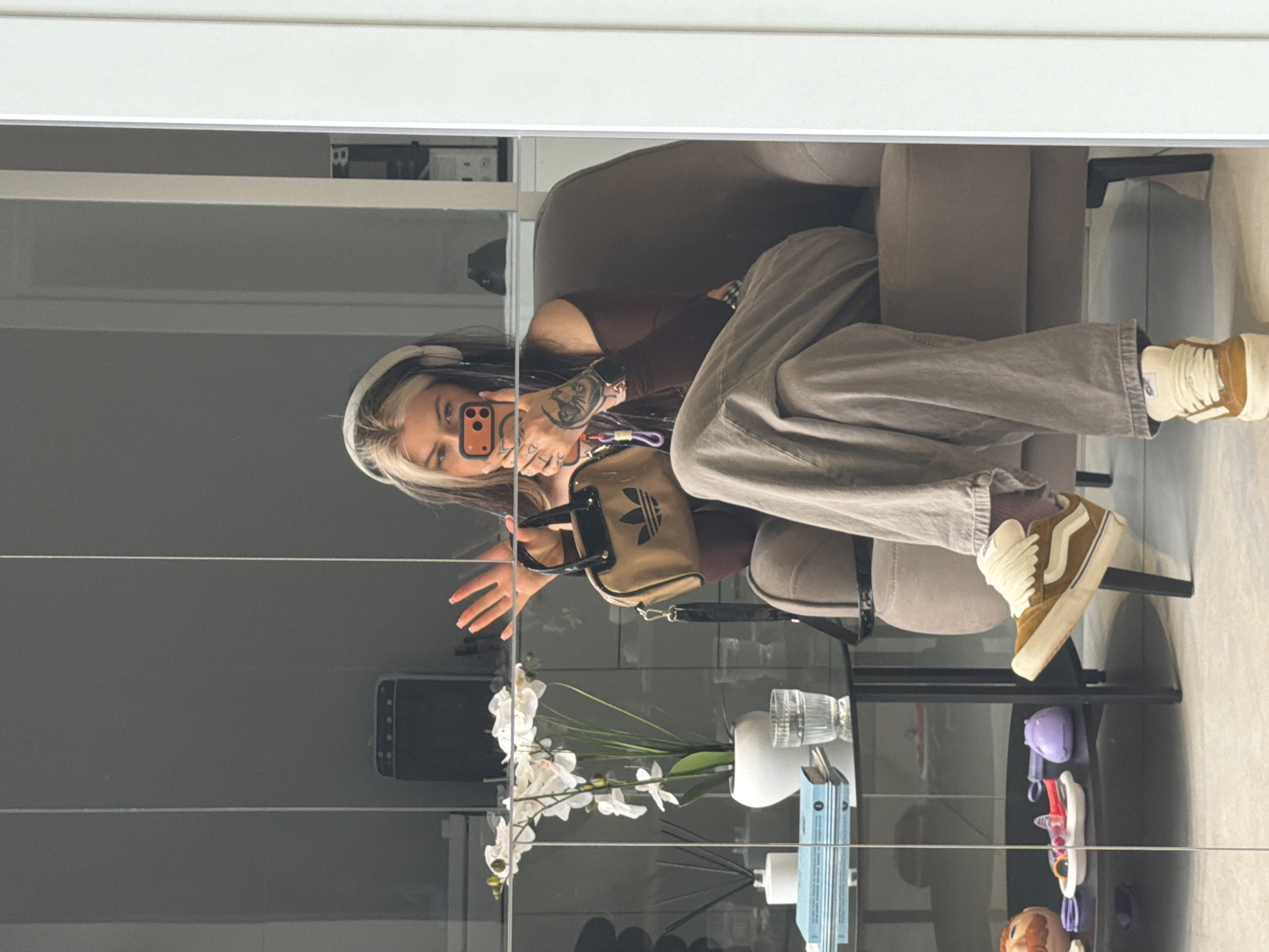 A young woman is captured in a mirror selfie, seated indoors. She is looking directly at the camera phone she holds with her right hand, which has a distinct orange case. Her left arm shows visible tattoos and she wears a smartwatch. She is dressed casually in a dark brown long-sleeved top and light grey baggy pants, paired with brown and white high-top sneakers. White headphones rest on her head, framing her hair which features contrasting dark and light (possibly blonde) streaks.
She is seated on what appears to be a light-colored armchair or sofa. To her left (viewer's right in the reflection), a dark-framed table holds decorative elements, including a white vase with white faux orchids, a clear glass, and a small blue box. On a lower shelf of the table, various small colorful items, including purple and white objects and a small figurine head, are visible. The background, reflected in the mirror, suggests an indoor setting, likely a room with light-colored walls and shelves. The small mini-bag she wears displays the "Adidas" logo. The setting implies a relaxed, personal moment, possibly in a home or hotel room in Budva, Montenegro.