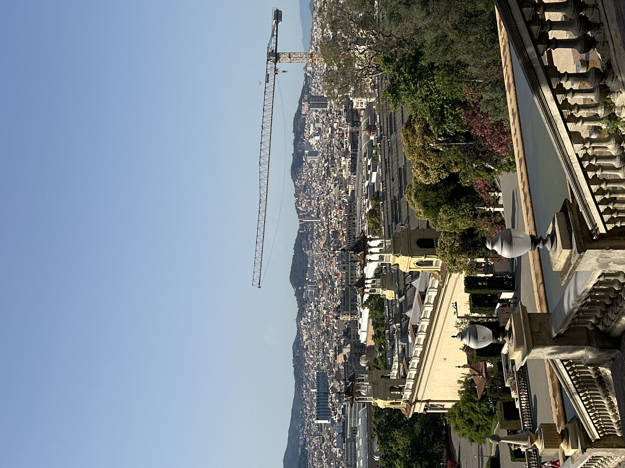This is an outdoor, eye-level, long-shot photograph of the city of Barcelona, Spain, taken from a slightly elevated position looking towards a dense urban landscape under a clear blue sky. In the foreground, a stone balustrade with ornate pillars and decorative lamps lines a walkway. Beyond this, a long, pale yellow building with multiple domes and turrets is visible, with a long rooftop extending backward. This building appears to be part of a historic complex.
The middle ground is dominated by a sprawling cityscape, characterized by a dense collection of buildings with red-tiled roofs and white facades. A construction crane, with its boom extended horizontally across the frame, is a prominent feature in the sky above the city. The background consists of rolling, green-covered hills that frame the city. Trees, some with dark green foliage and others with vibrant pink blossoms, are scattered throughout the scene, particularly around the historic building. A few people are visible, small figures on the walkway in the foreground, appearing to be pedestrians. The overall impression is one of a sunny day in a historically rich, yet developing, urban environment.