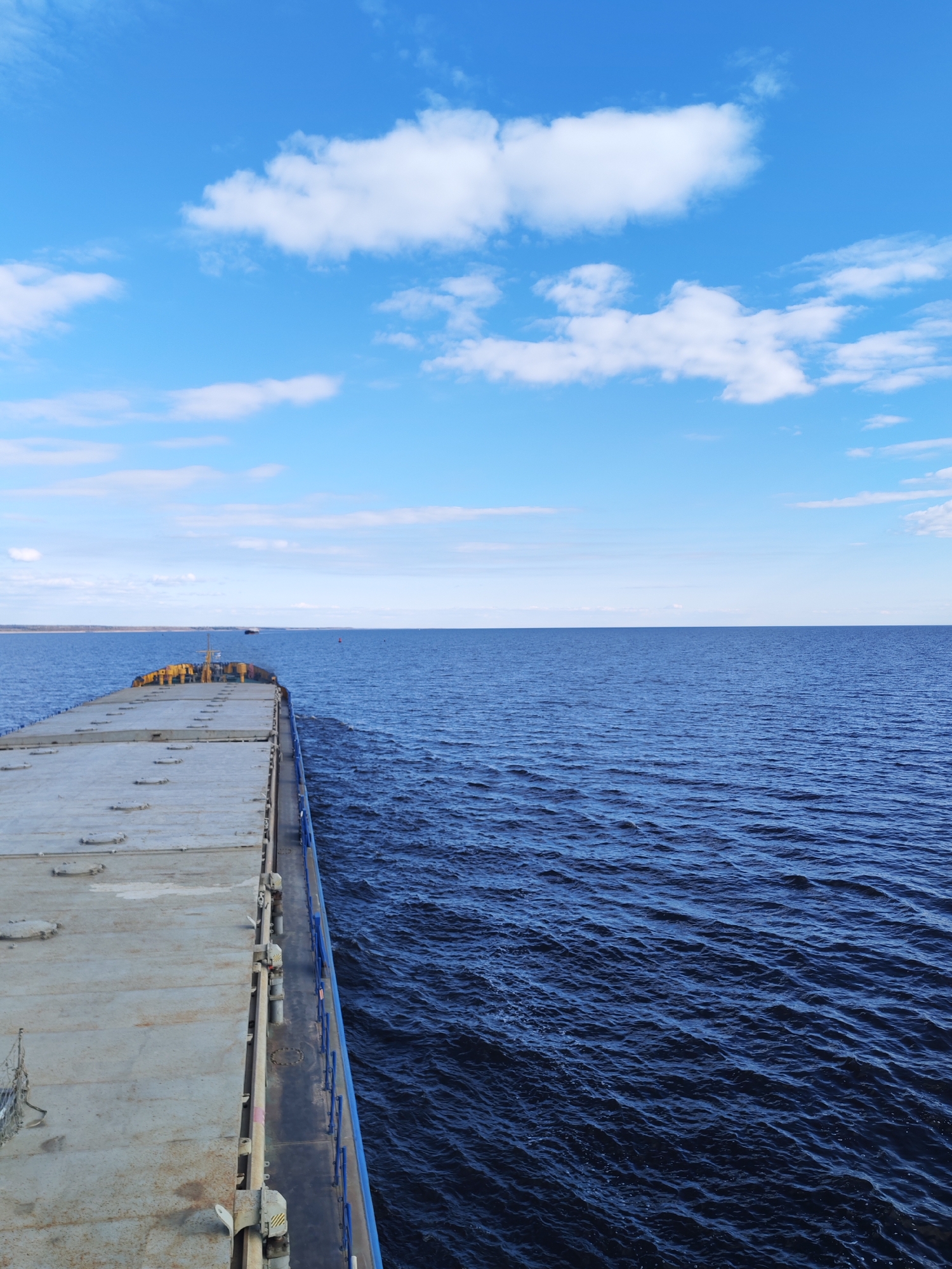 The image presents a broad daytime view from the deck of a large cargo vessel, likely a barge or bulk carrier, navigating a wide body of water. The ship's flat, utilitarian deck, fitted with numerous sealed hatches, occupies the foreground and extends into the middle distance on the left. A blue railing runs along the starboard side of the vessel, bordering the dark blue water.
The water itself is a deep, rich blue, showing gentle ripples and small waves across its surface, suggesting calm to moderate conditions. A faint wake or disturbance in the water along the ship's side indicates that the vessel is actively moving forward. On the distant horizon, a low-lying, light brown strip of land stretches across the frame, implying a large river or lake rather than open ocean. A small, lighter-colored vessel is barely discernible on the horizon near the distant shoreline.
The sky is a clear and vibrant blue, adorned with scattered white cumulus clouds of various sizes, typical of fair weather. The bright and even illumination suggests it is mid-day. The scene evokes a sense of vastness and tranquil travel.
No people are visible in the image. There is no discernible text or specific markings on the ship or in the environment. The setting, with its large waterway and distant low land, is consistent with the geographical context of Sannikovo, Russia, which is situated near major river systems.