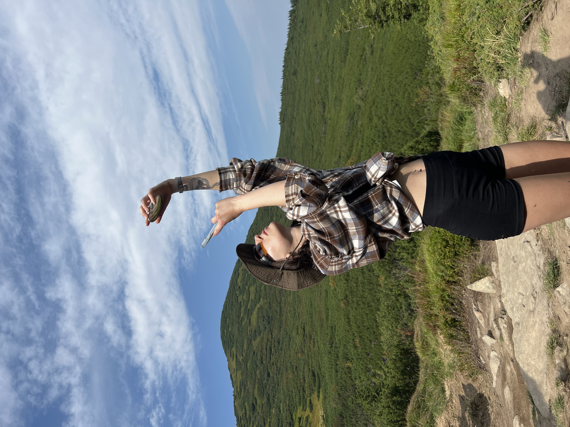 A person wearing a brown plaid shirt, black shorts, sunglasses, and a brown hat stands on a rocky path in a mountainous area. The person is holding a smartphone up towards the sky with their right hand, and their left hand is also raised, holding a small circular object. The person has a tattoo of a snake on their left arm and a belly button ring. The background features a lush green mountain covered in trees and brush, under a blue sky with scattered white clouds. This scene is likely located in or near the Carpathian Mountains, which are a prominent geographical feature in the Ivano-Frankivsk Oblast region of Ukraine, where the city of Vorokhta is situated.