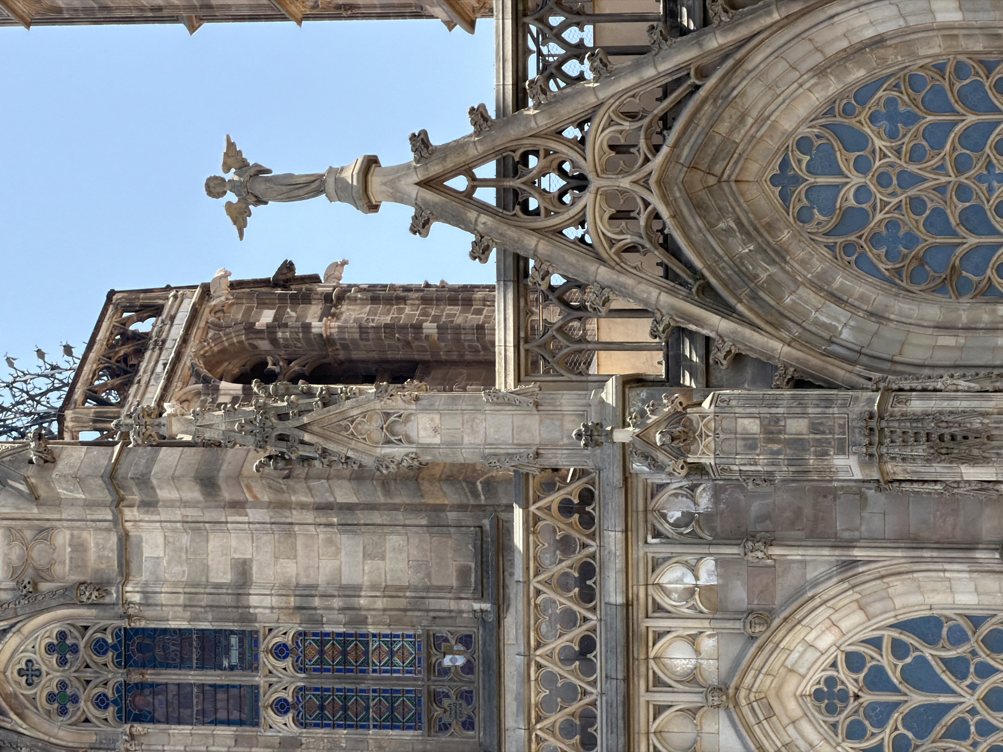 The image shows a detailed view of the exterior of the Barcelona Cathedral, located in the Gothic Quarter of Barcelona, Spain. The photograph focuses on the intricate Gothic architecture, particularly the stonework, stained glass windows, and decorative elements. A statue of an angel with outstretched wings is visible atop a pillar on the right side of the frame, set against a clear blue sky. The facade features pointed arches, flying buttresses, and ornate tracery. The stained glass windows display geometric patterns and figures in shades of blue, green, and red. The stonework shows signs of age and weathering, with visible mortar lines and variations in the color of the stone. The overall scene conveys a sense of historical grandeur and architectural complexity.