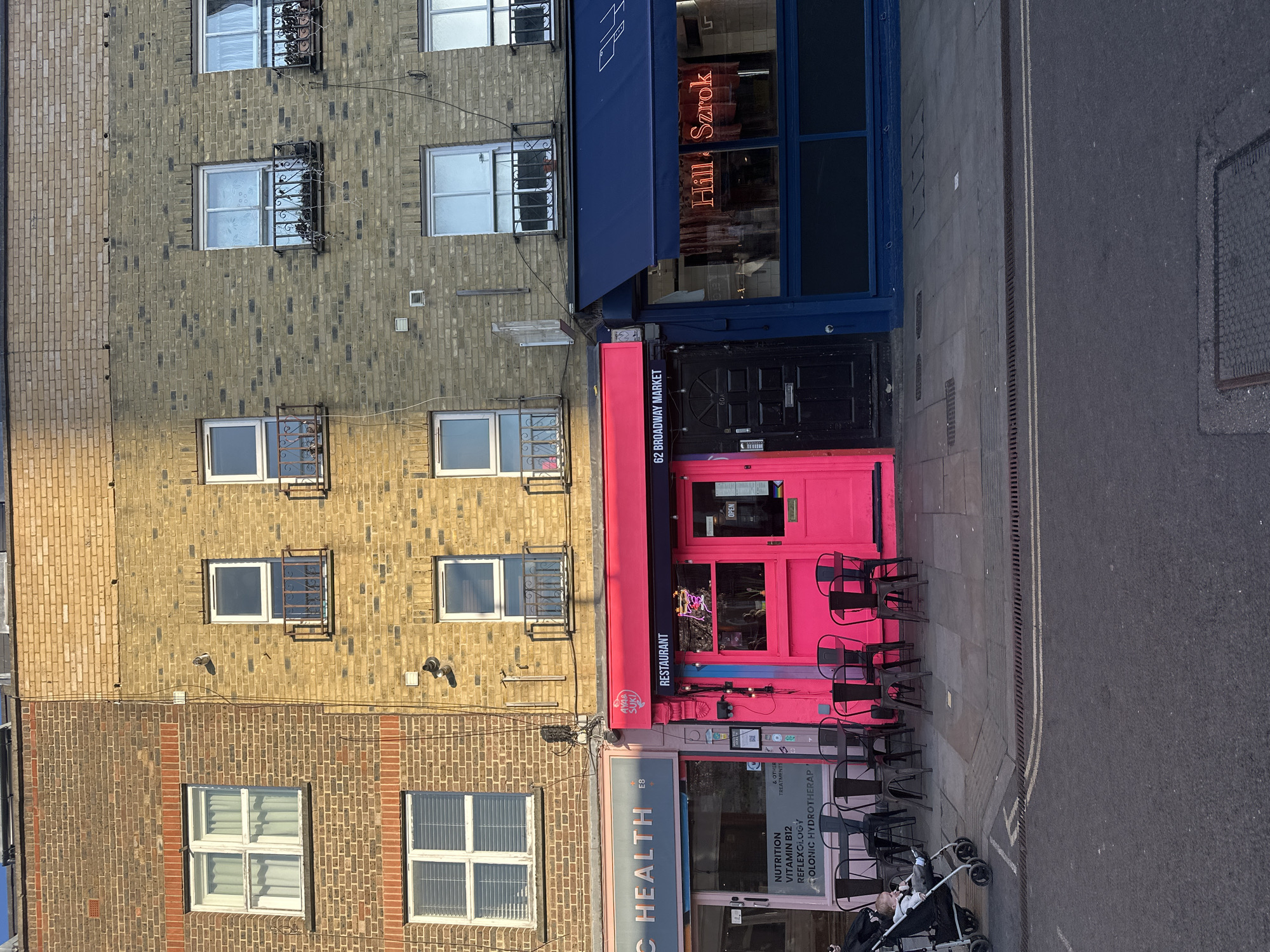 The image shows a street scene in London, United Kingdom, featuring several storefronts.
The store at 62 Broadway Market is painted a vibrant pink and is labeled as a restaurant. Next to it is a blue storefront. Adjacent to the restaurant is a health clinic or wellness center with text visible stating "NUTRITION VITAMIN B12 REFLEXOLOGY." A stroller is visible in front of the health clinic.
The buildings are brick and show signs of aging. The street is gray and appears to be a typical London street.