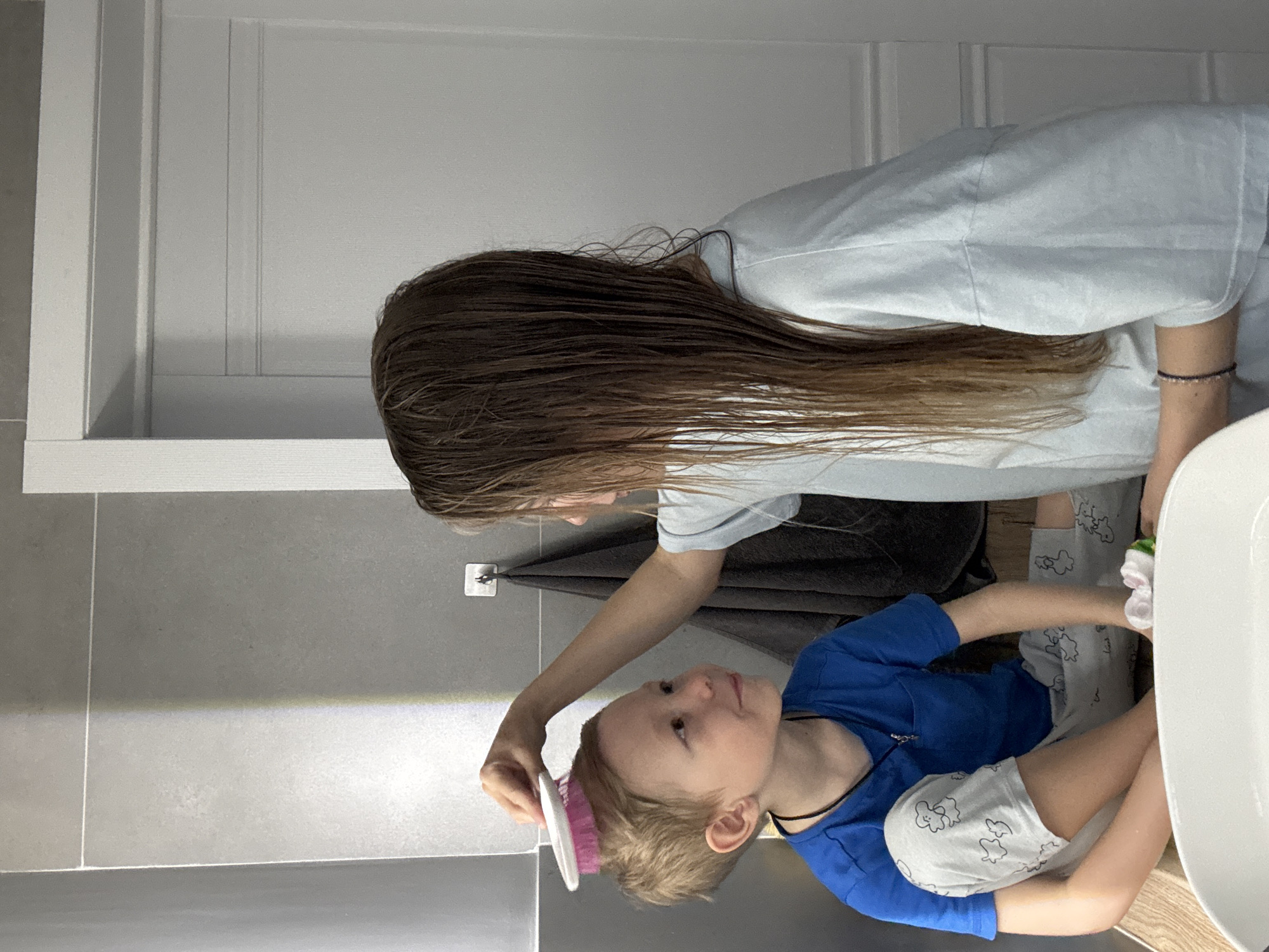 A woman is brushing a young boy's hair in a bathroom. The boy is wearing a blue shirt and patterned grey shorts. The woman has long, wet brown hair and is wearing a light blue shirt. A dark grey towel hangs on the wall behind them. The bathroom has grey and white tiled walls. The location may be in Chaikovsky, Russia.