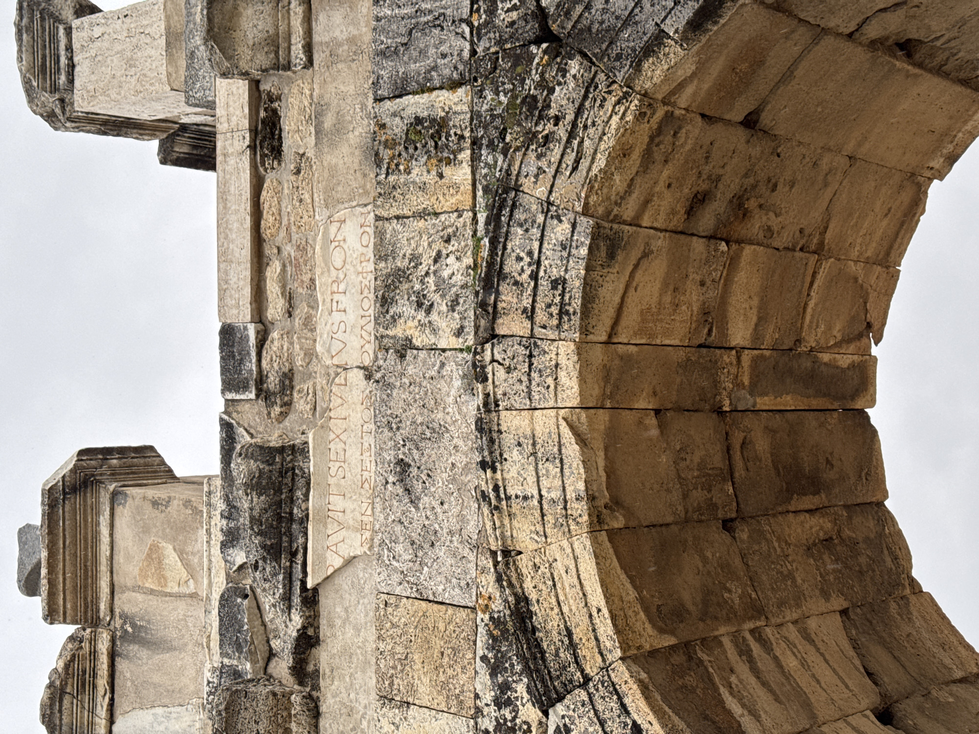 The image shows an archway constructed of stone blocks, likely part of an ancient structure. The location is Pamukkale, Turkey, based on the presence of identifiable ancient Roman ruins. The archway is composed of carefully fitted stone blocks, some of which have weathered over time, with visible lichens and discoloration. There is an inscription on a section of the arch. The inscription is in Latin. The background is a plain sky.